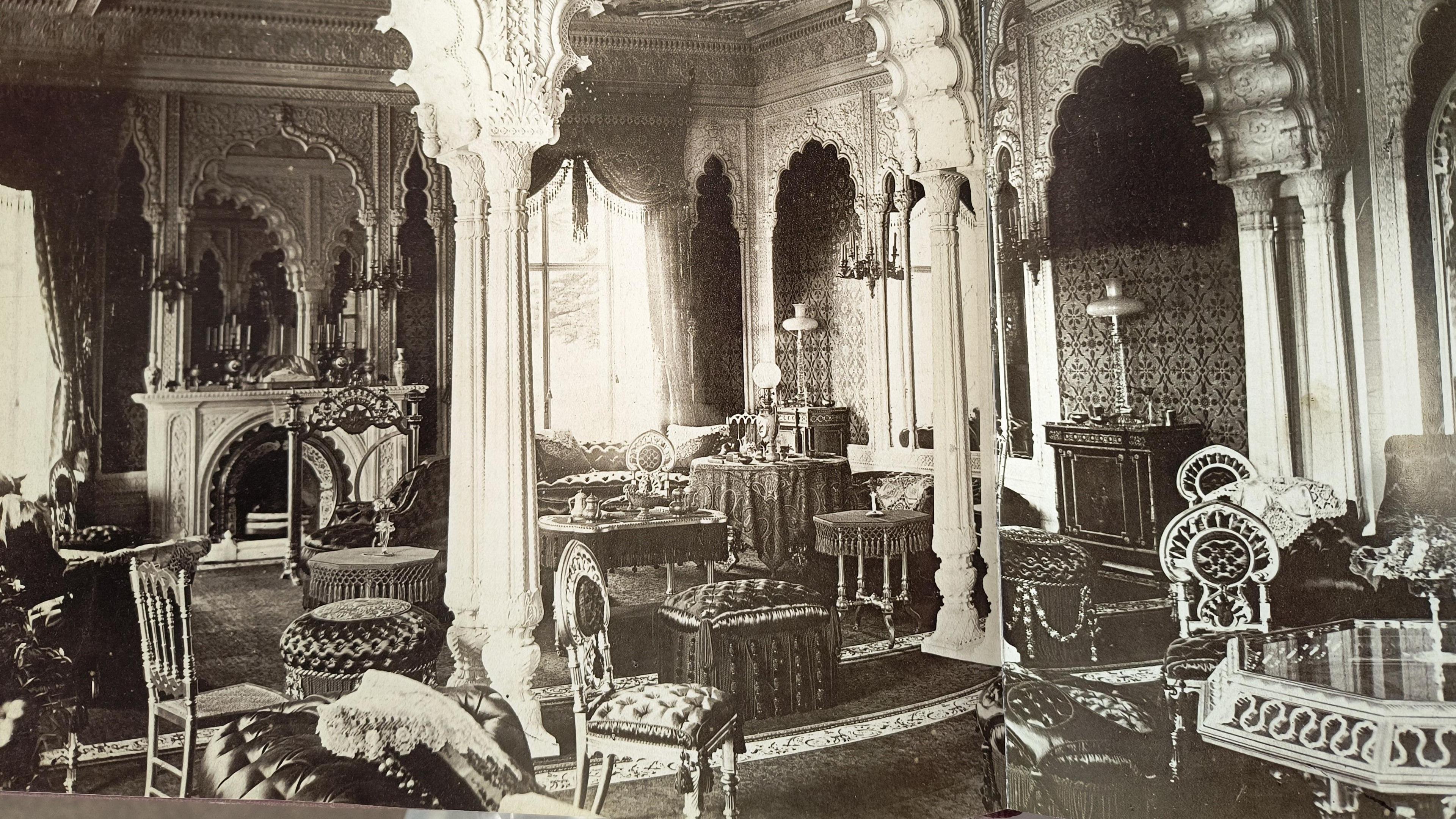 A black and white image showing the inside of Elveden Hall. There are a number of tables and ornate chairs spread out in a room with a large fireplace on one side, and mirrored walls on the other. 