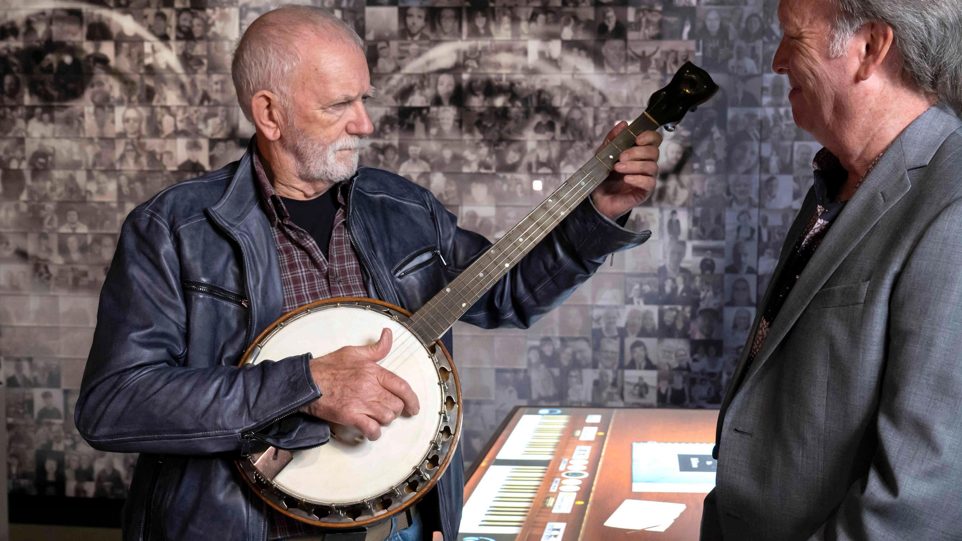 Rod Davis, with a balding head and grey beard, wears a blue leather jacket and mauve and grey checked shirt and holds a white and black banjo in front of a wall of photographs which form a collage of John Lennon's bespectacled face