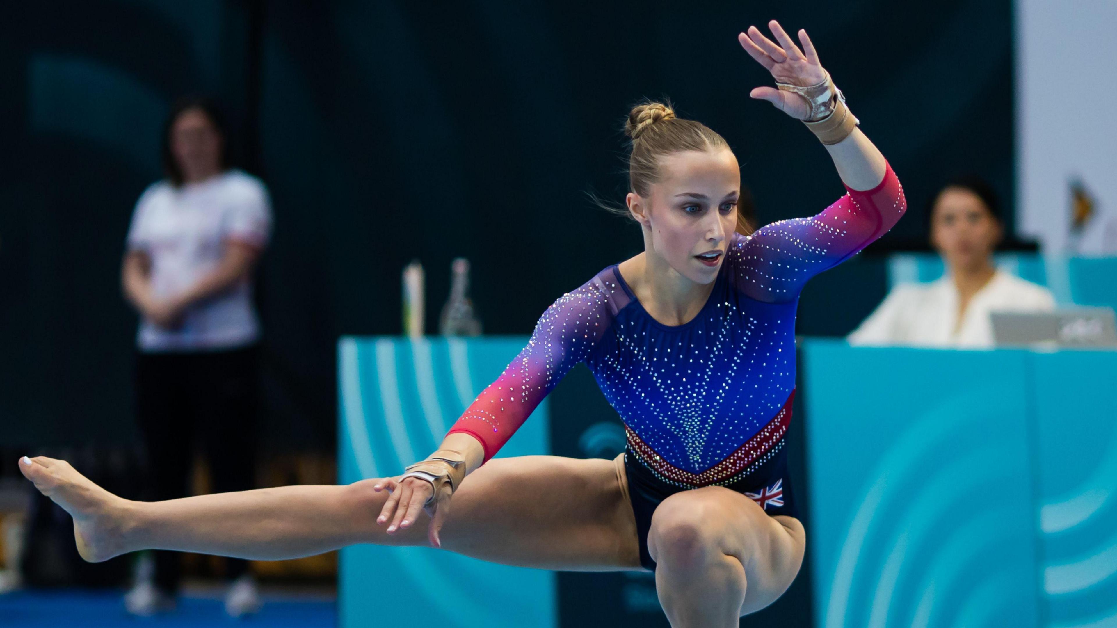 Abigail Roper during a floor routine. She is wearing a sparkly blue and red leotard. One leg is planted on the floor and the other is held outwards at a 90-degree angle to her body. She has a hand raised above her head.