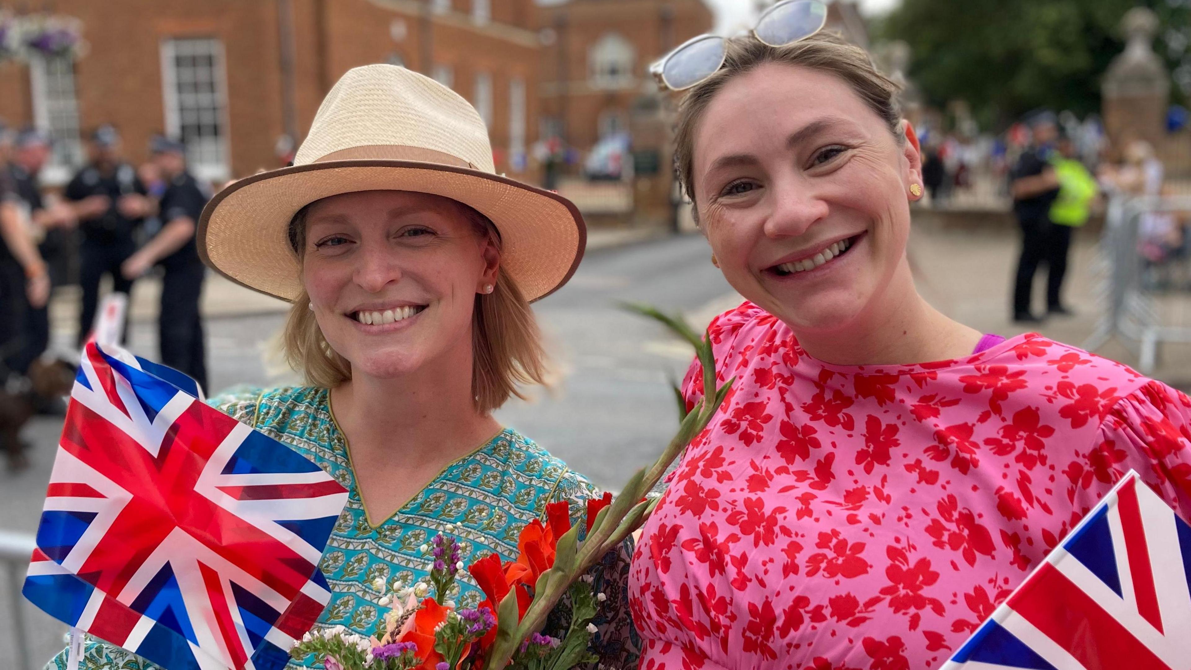 Bronwyn and Bella Iten-Scott smile at the camera on Newmarket High Street. Bronwyn wears a hat and green dress and she holds a Union Jack flag with flowers. Bella has sunglasses on her head, wears a pink dress and holds her flag. 