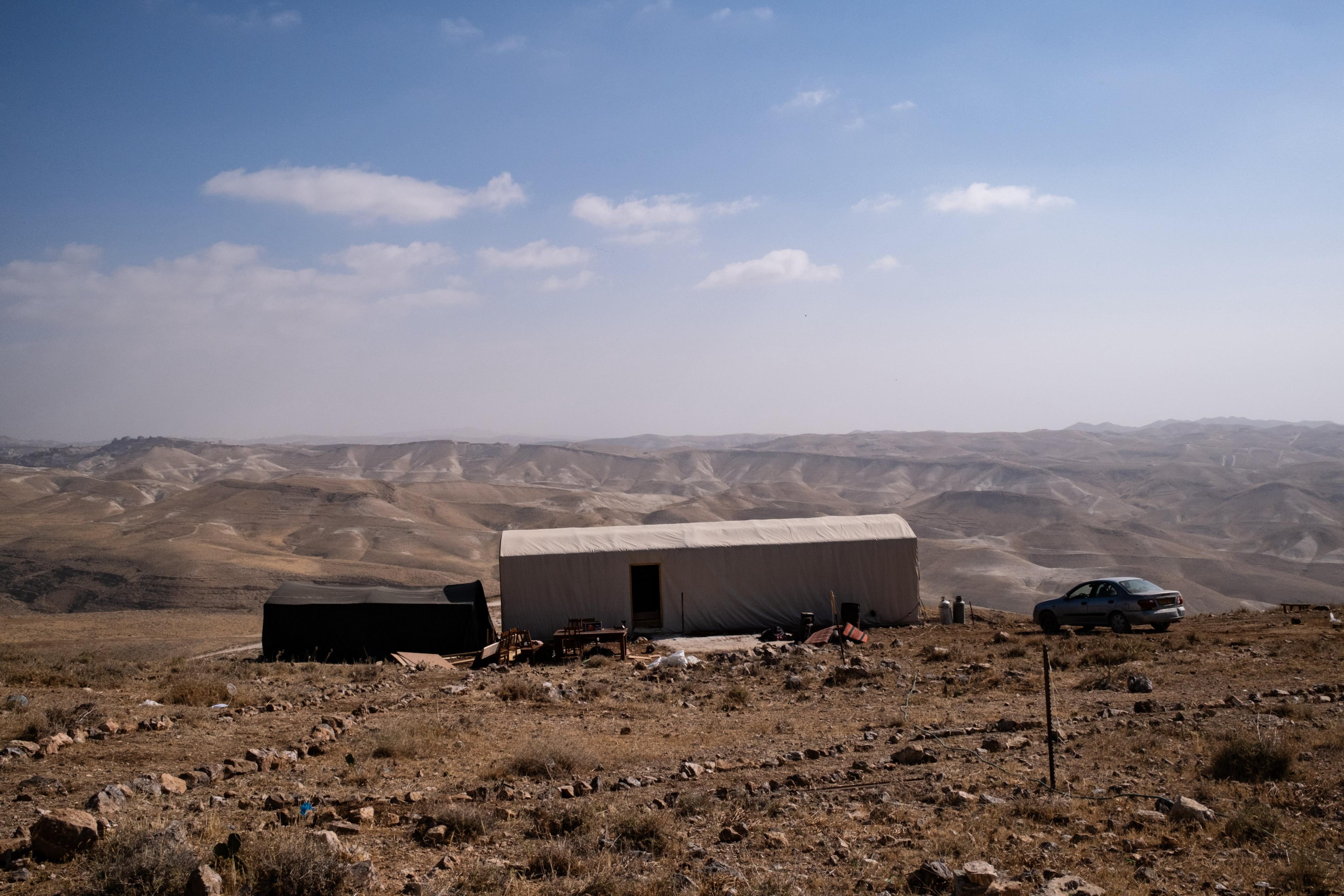 An outpost in the West Bank - beige non-descript building and a car overlook a valley with mountains in the distance. The sky is blue with some small white clouds. 