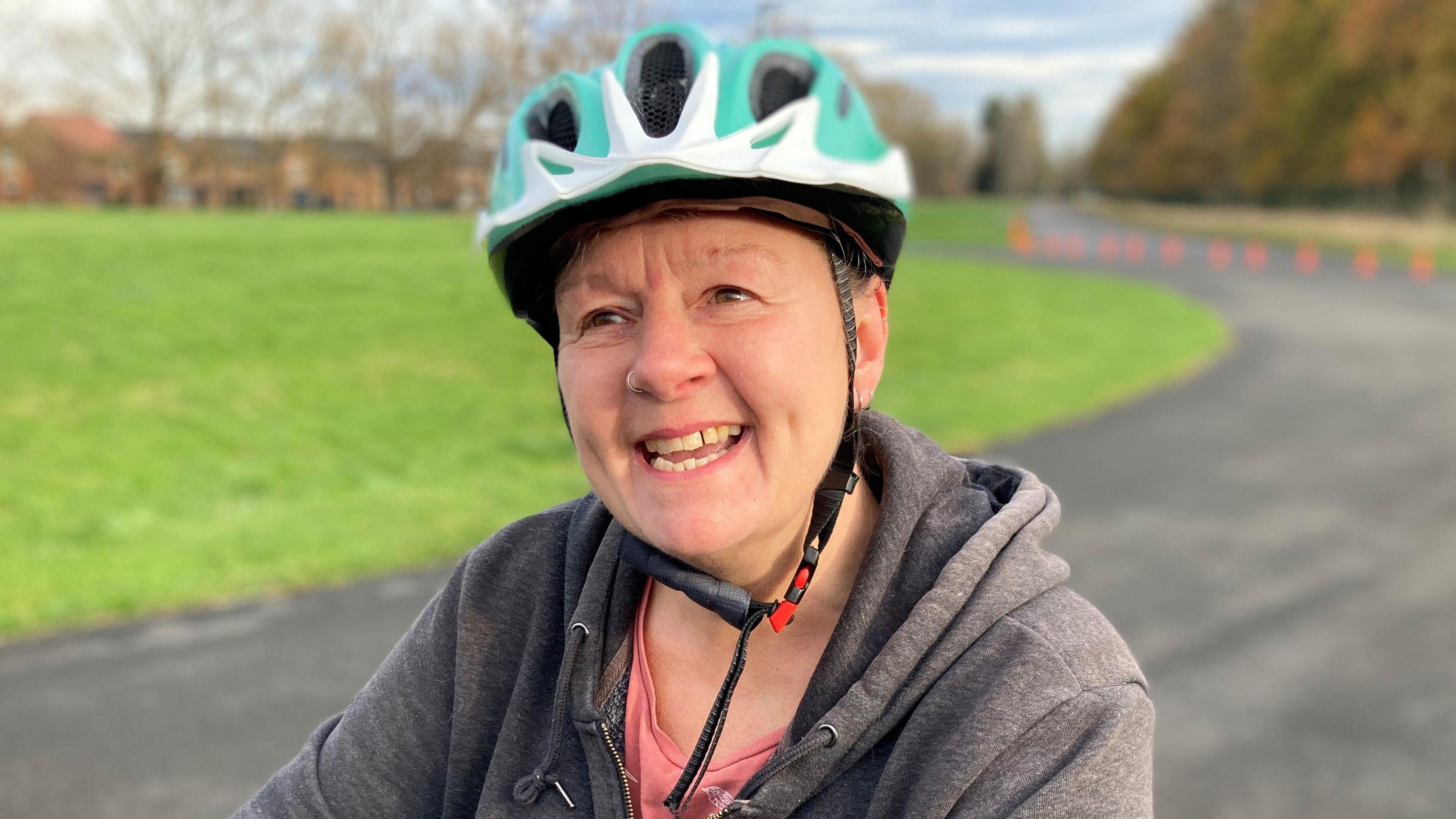 Sarah Smith is on her bike, on a cycle track, and is smiling. She is wearing a green and white helmet and a grey jumper.