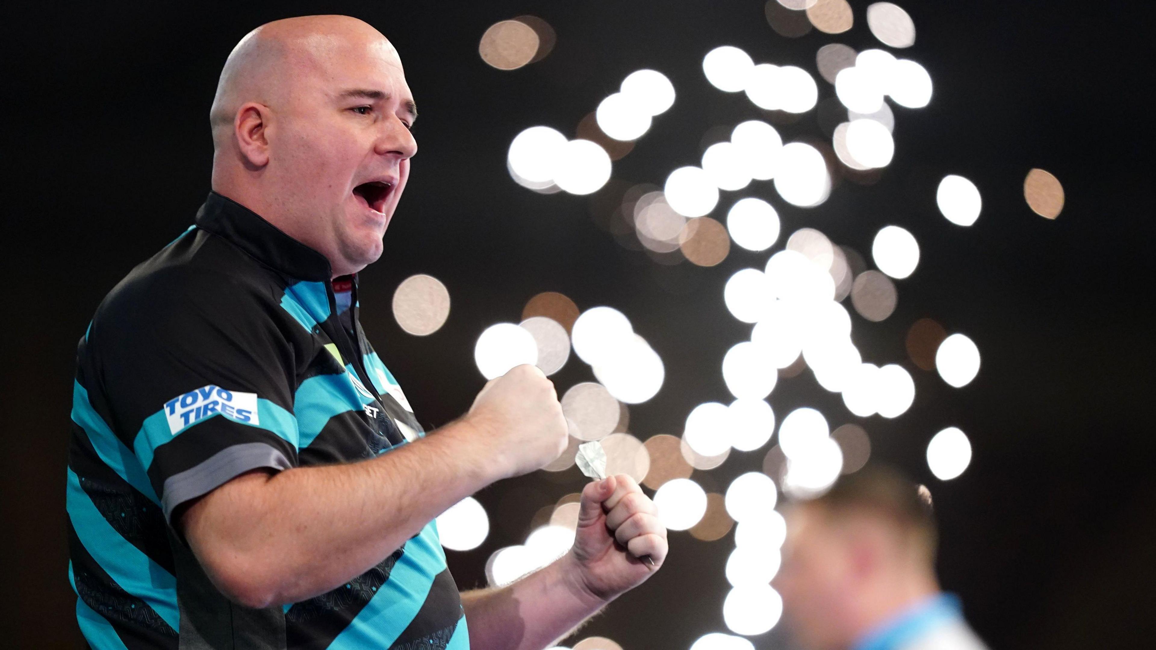 Rob Cross celebrates after beating Chris Dobey on day fourteen of the Paddy Power World Darts Championship at Alexandra Palace, London. He is wearing a blue and black shirt and is clenching his fists. 