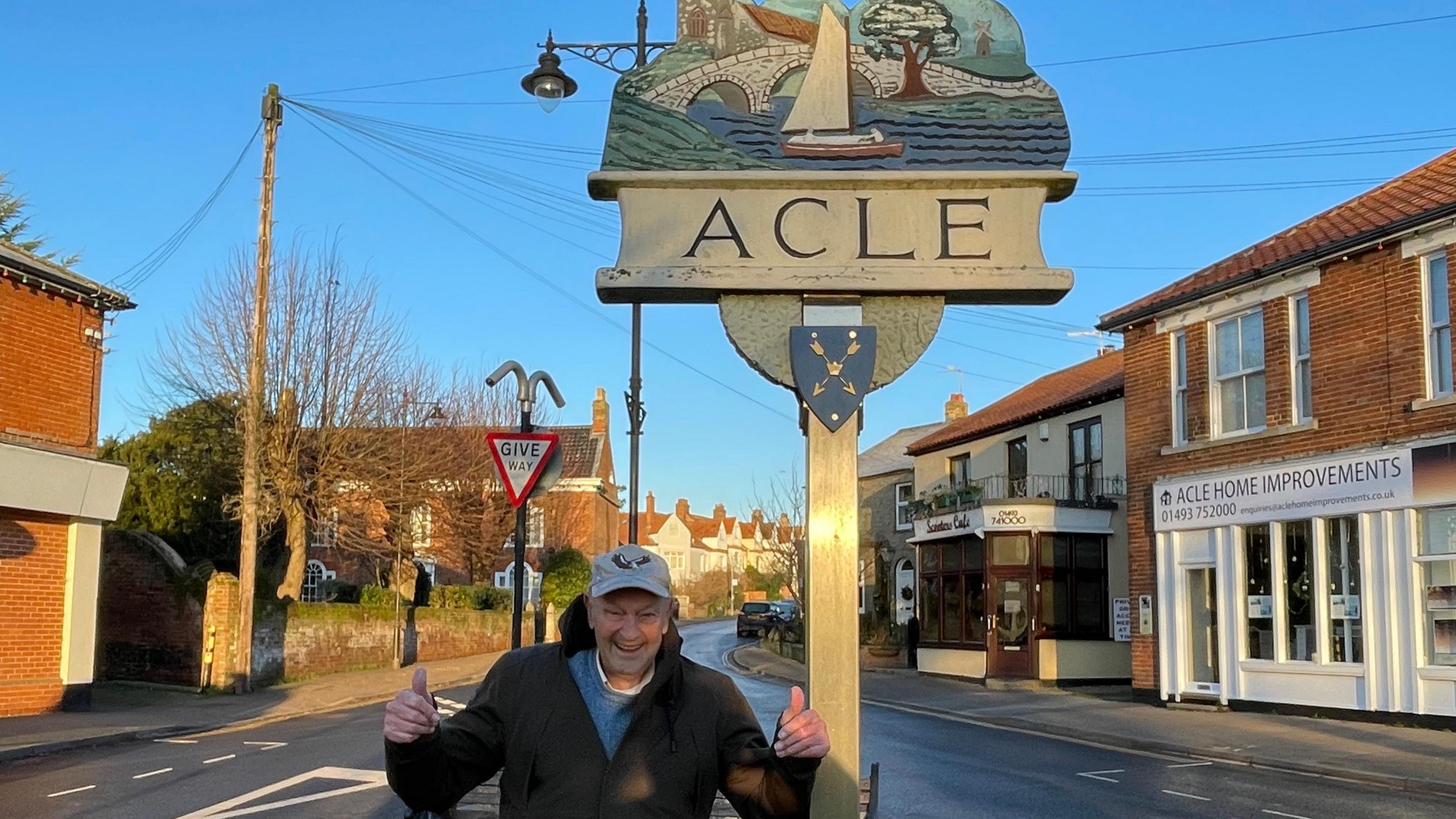 Malcolm Metcalf next to the Acle village sign wearing a black or dark-coloured coat. A few shops can be seen to the right of the frame.