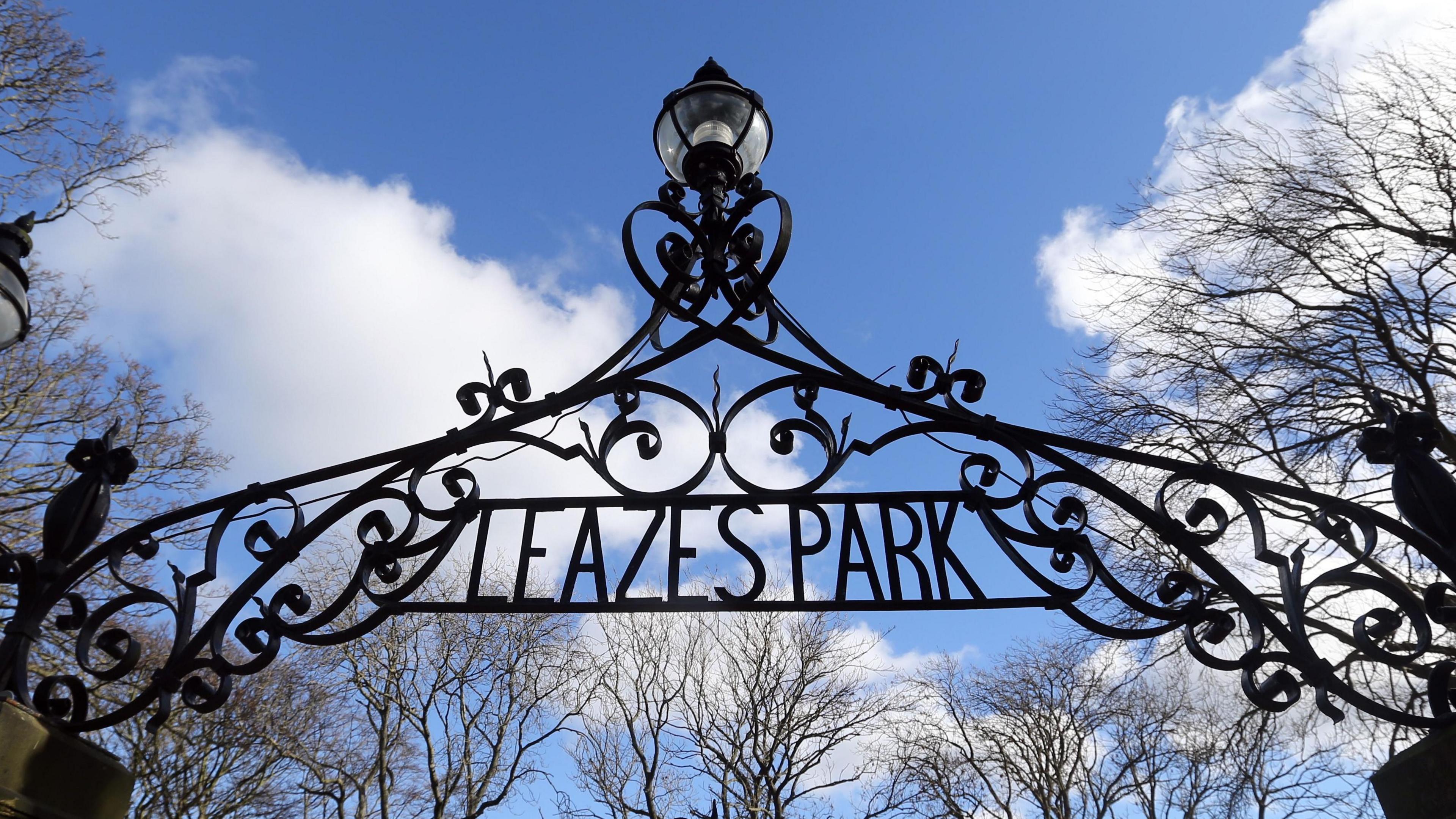The metal arch above the entrance to Leazes Park. Black metal is bent into a swirling design, which is topped with a lamp and contains the name of the park.