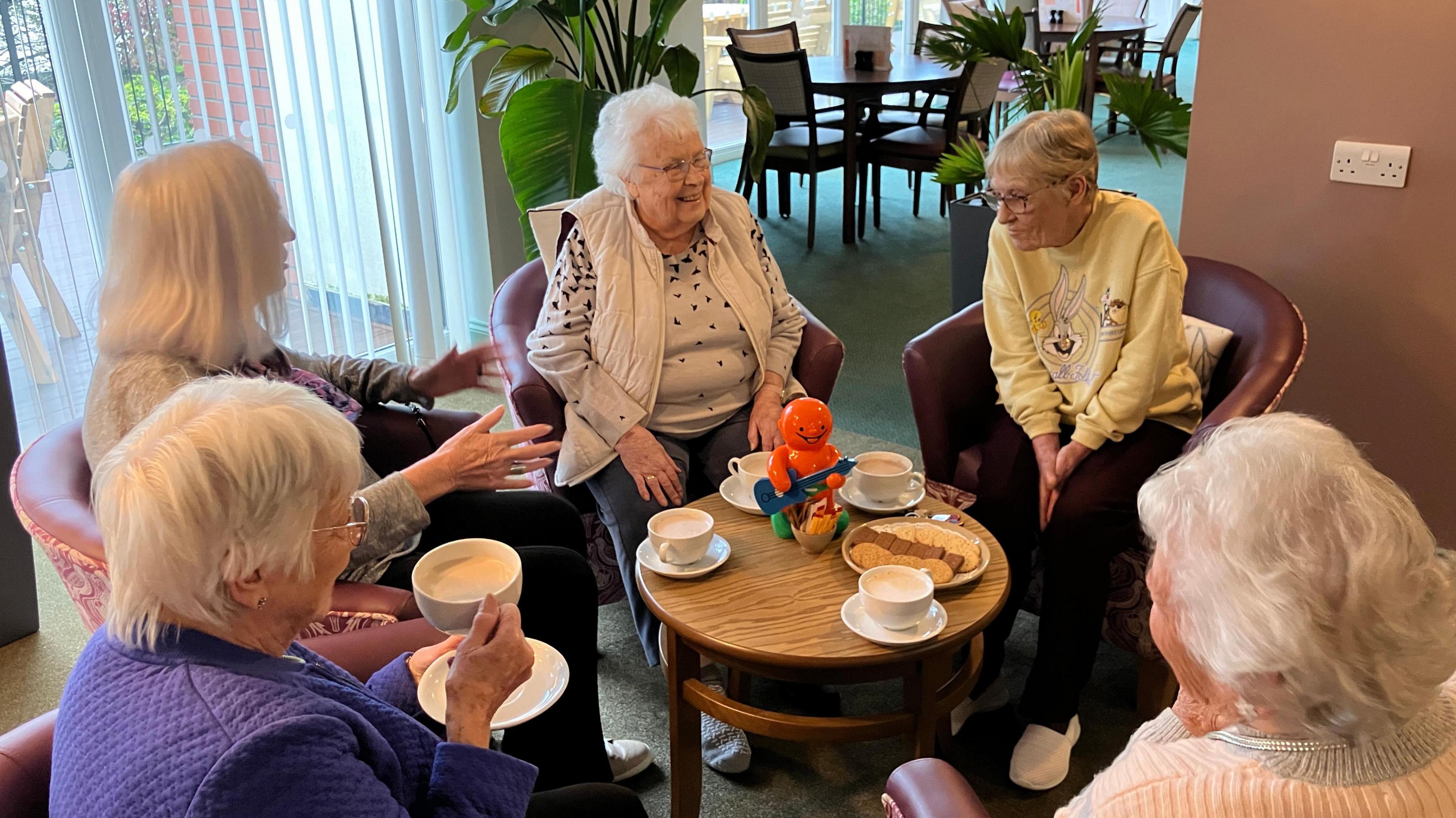 Monica Wingrove sits in a circle surrounded by friends drinking coffee and easting biscuits. On the table is a red collecting tin in the shape of a jelly baby. 