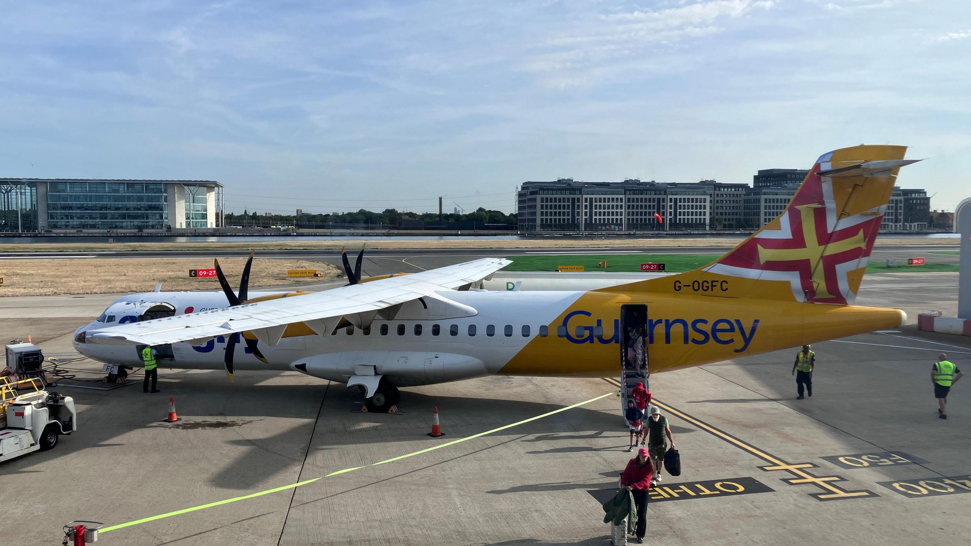 An Aurigny flight on the runways of an airport. People are boarding the plane from the rear. Building are in the distance and it's a sunny day with blue sky and a few clouds