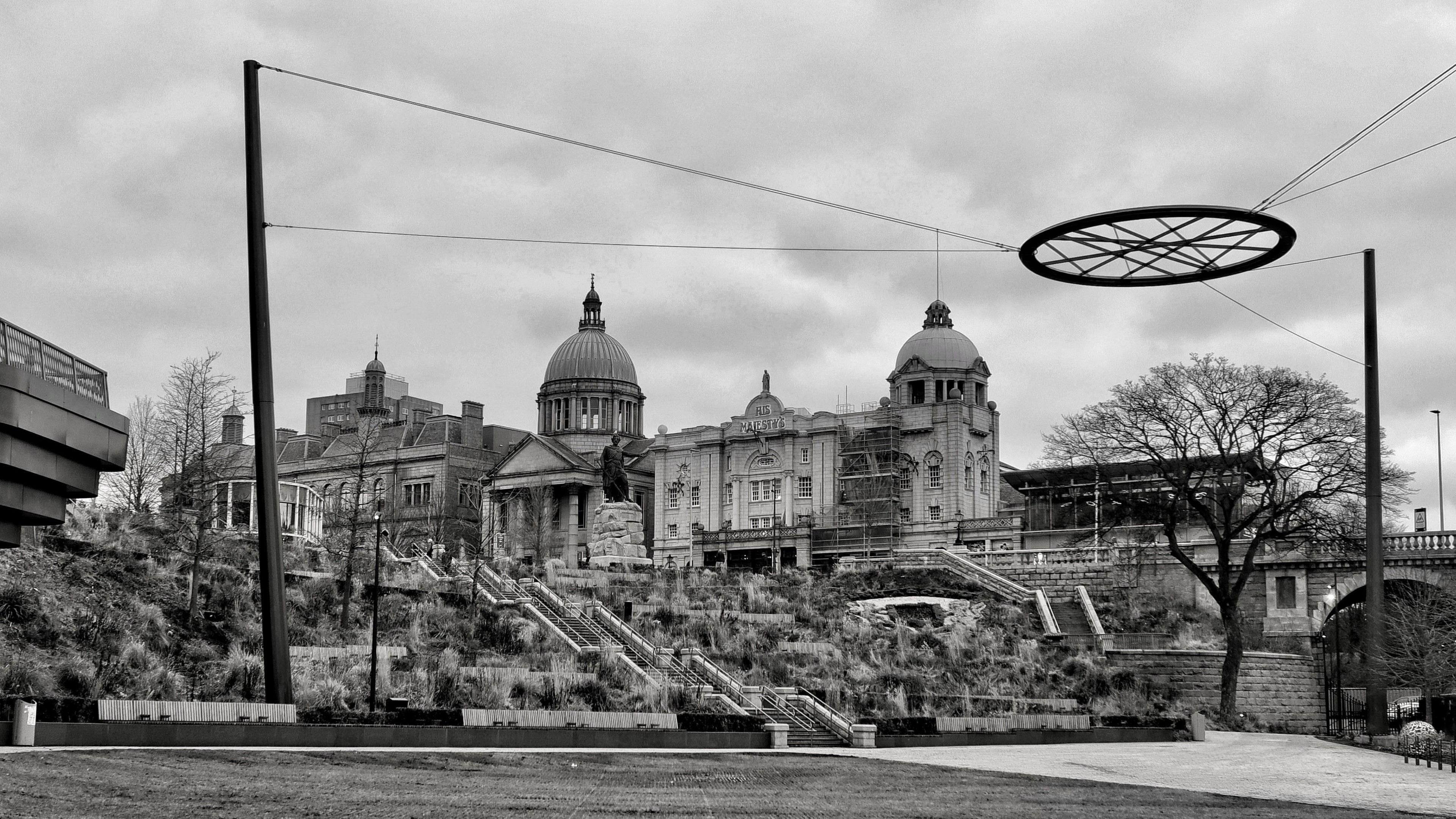 A circle structure hangs from ropes above a park. An ornate building with domes is in the background. It is a black and white picture.