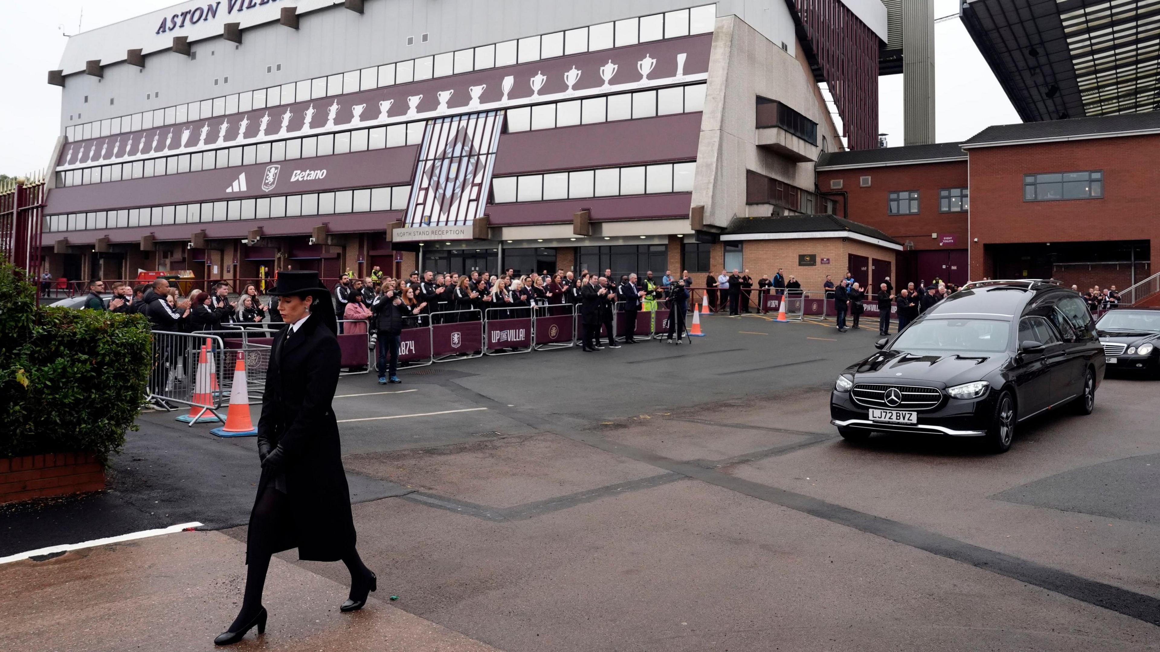 A funeral procession, led by a woman wearing a black hat with a veil down the back,  makes its way past Villa Park with crowds gathered behind a barrier outside the stadium. The woman is followed by two black cars.