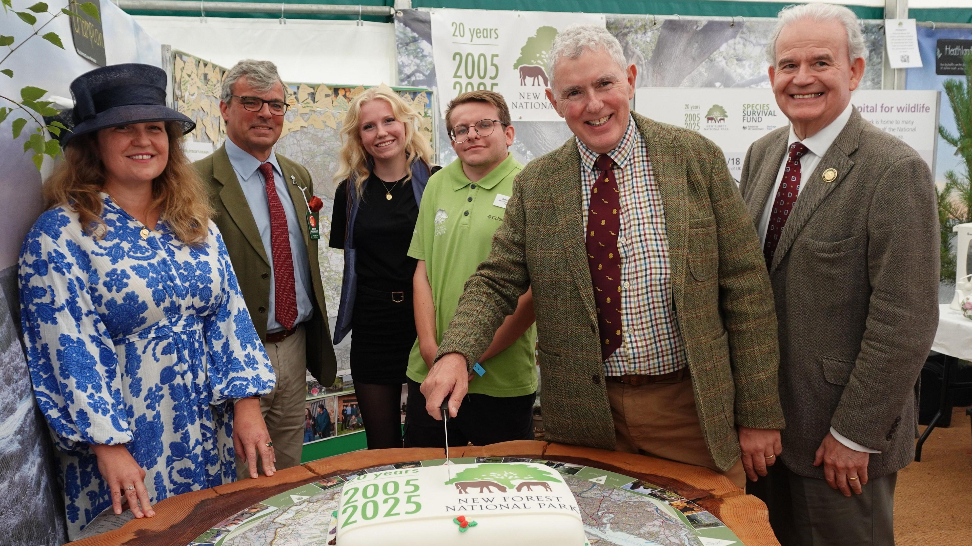 A group of four men and two women posing next toa large square cake decorated with 2005 to 2025 and the New Forest National Park logo of a green tree and two brown ponies. Three of the men have grey hair and are wearing tweed jackets and maroon ties. One of the women is dressed in a blue and white patterned dress and a large blue wedding-style hat. The other man and woman are dressed casually.
