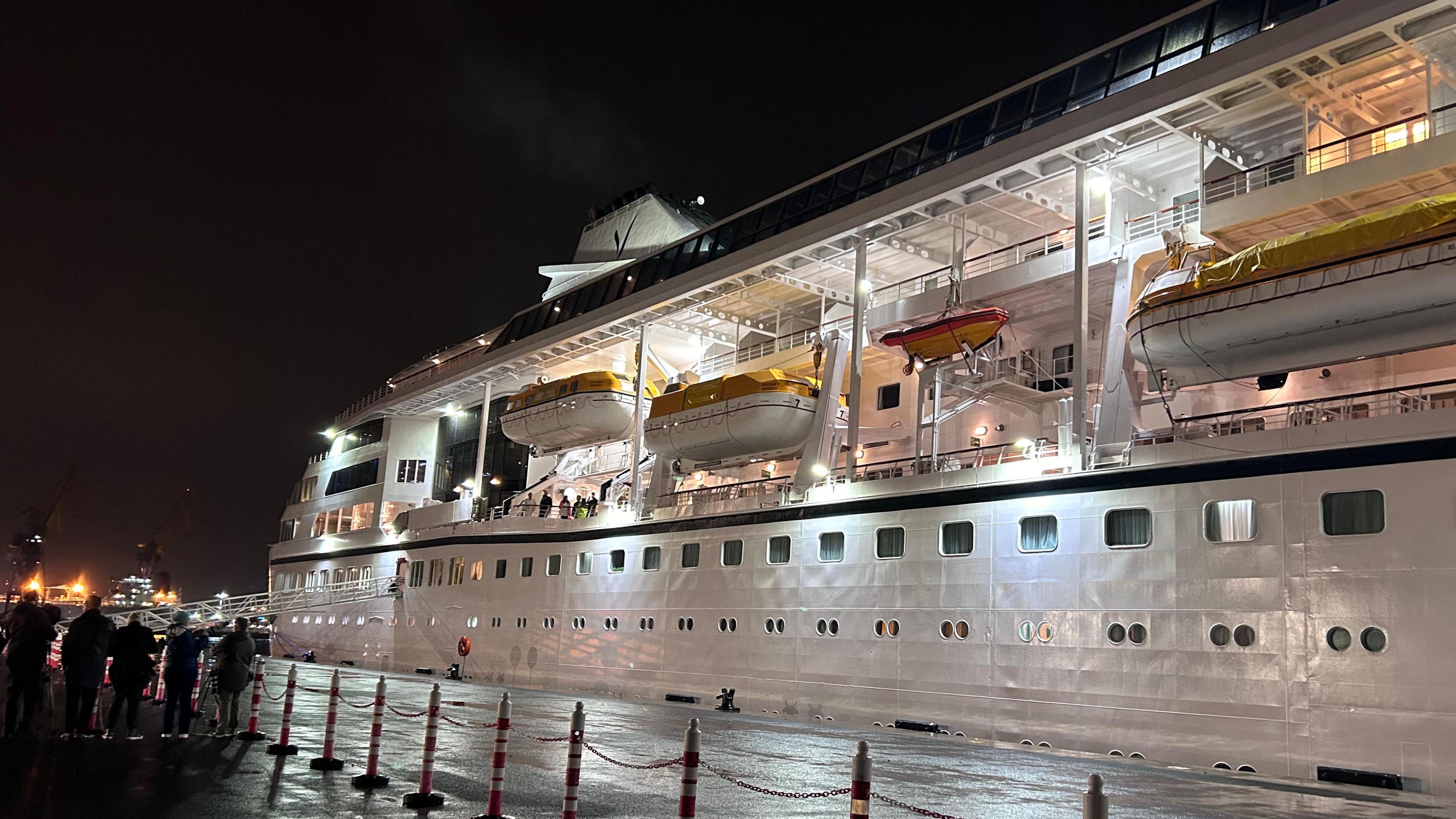 The ship has three stories and lots of small windows.

The sky is black and there are lights on in the windows. 

Passengers are visible on the ship. 