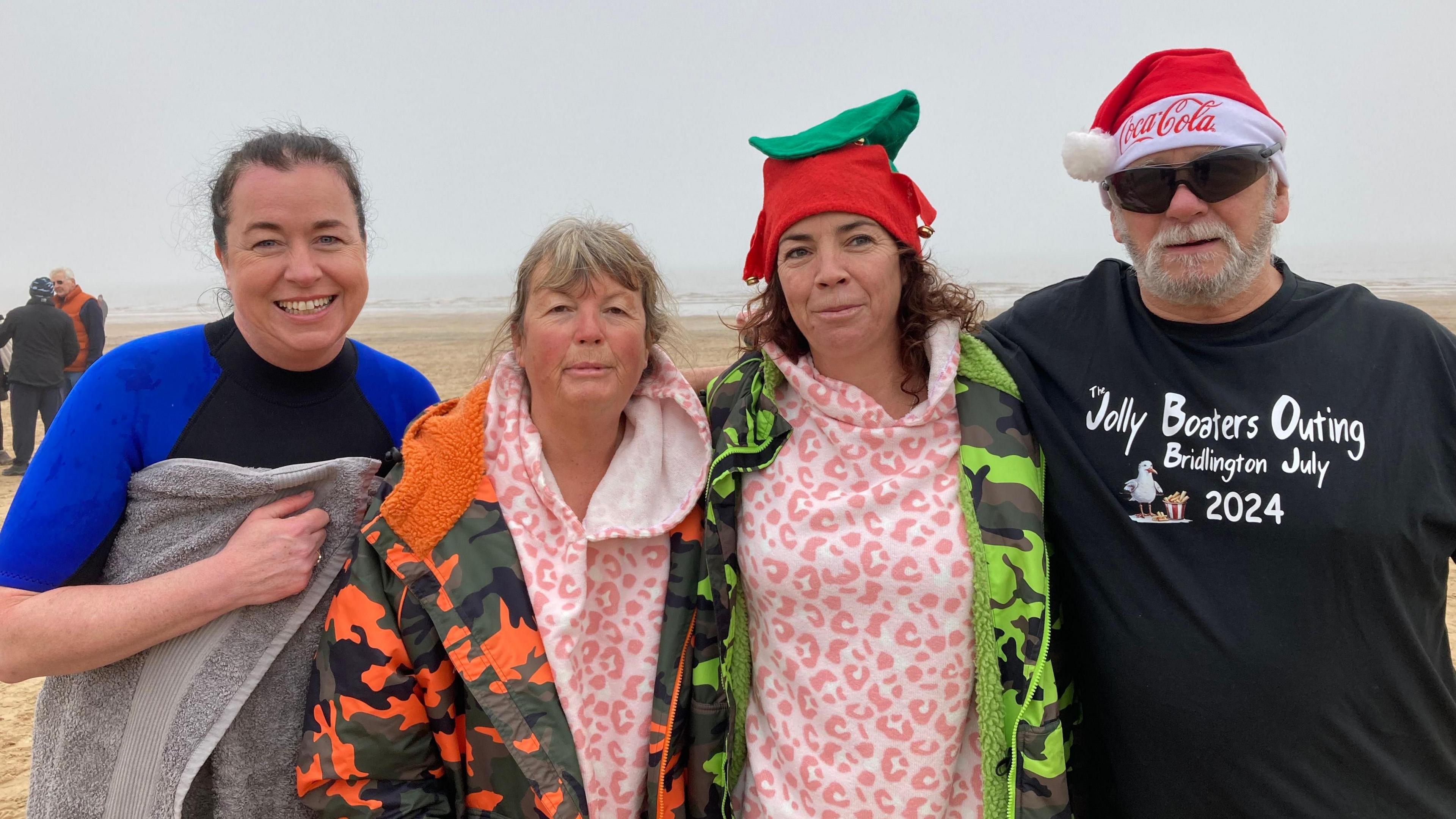 Four people looking into the camera and smiling. From left to right: A woman with tied-back brown hair wearing a blue and black swimsuit with a grey towel wrapped around her; a woman with light-coloured hair wearing a pink cow-print jumper and an orange camouflaged coat; a woman with brown hair wearing a pink cow-print jumper, a green camouflaged coat and a red and green elf hat; and a man wearing a black t-shirt that reads "The Jolly Boaters Outing. Bridlington July 2024". He is also wearing black sunglasses and a red and white Santa hat. 