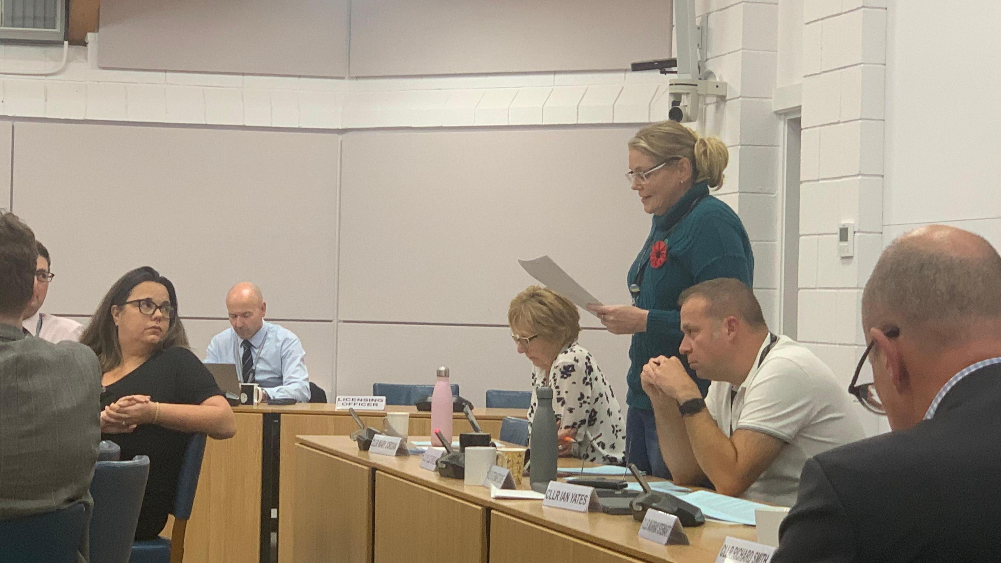 Liz Skelt stands up at her desk at a council meeting and reads off a piece of paper. She is wearing a long-sleeved emerald dress and a Remembrance Day poppy, and has her blonde hair in a ponytail. She is also wearing glasses. Her fellow councillors are listening, with one female councillor sat in front of her turned around in her seat.