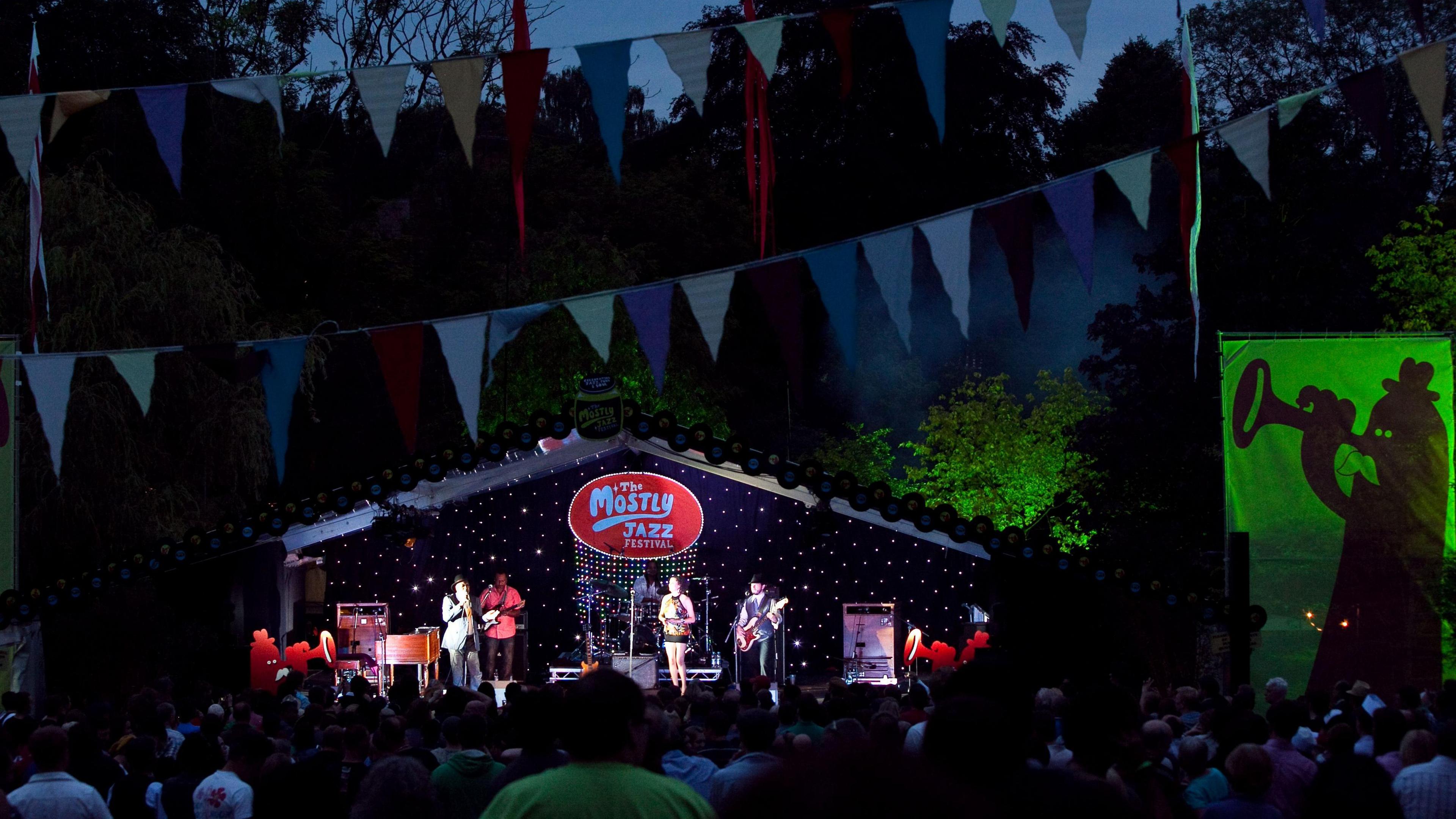 Artists are standing on a stage with a sign that says "The Mostly Jazz Festival". Colourful bunting is strung through the trees.