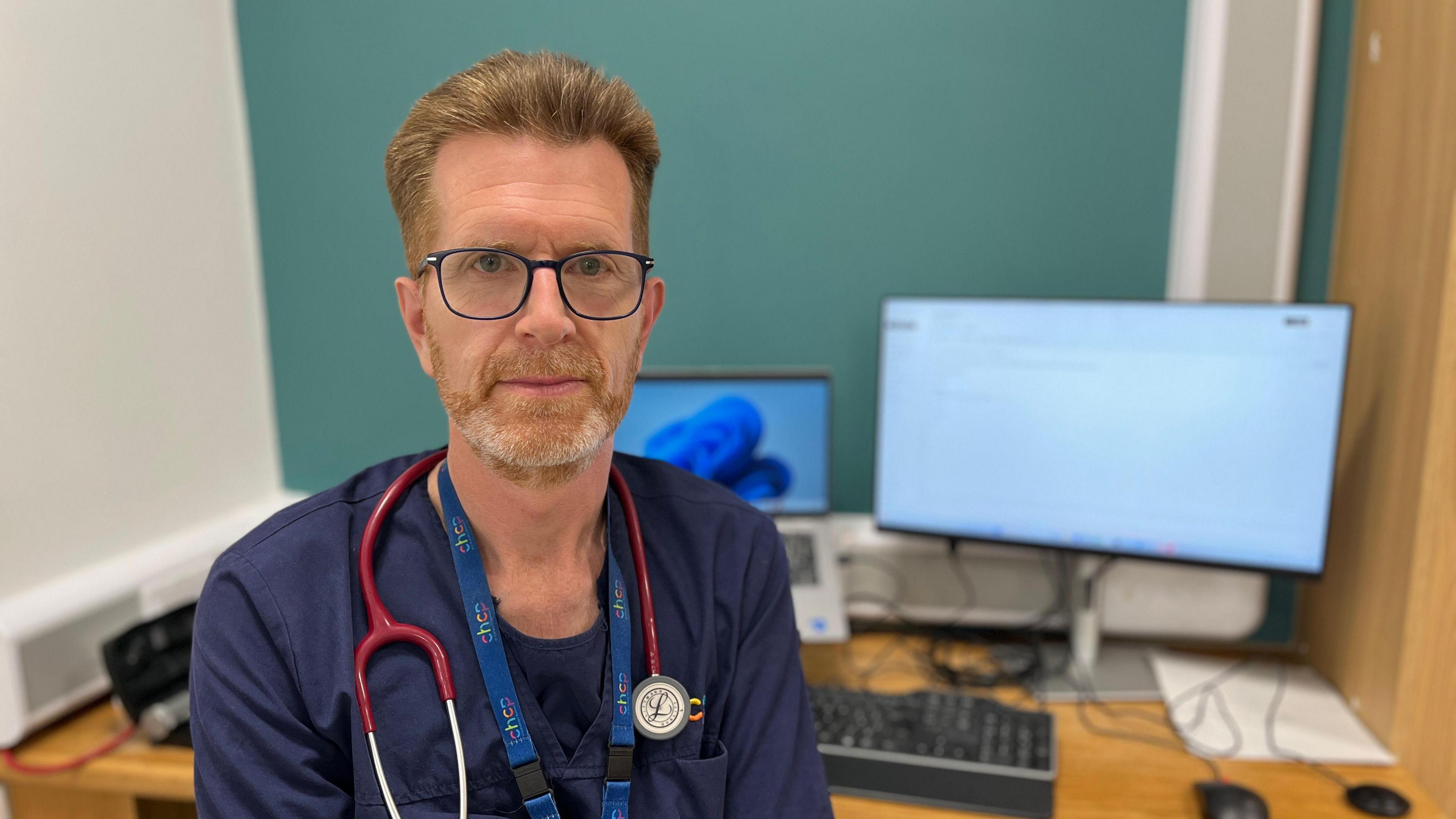 A man with short, strawberry blond hair and a beard and moustache. He is wearing dark-rimmed glasses and has a stethoscope around his neck. He is sitting in front of two computer screens.