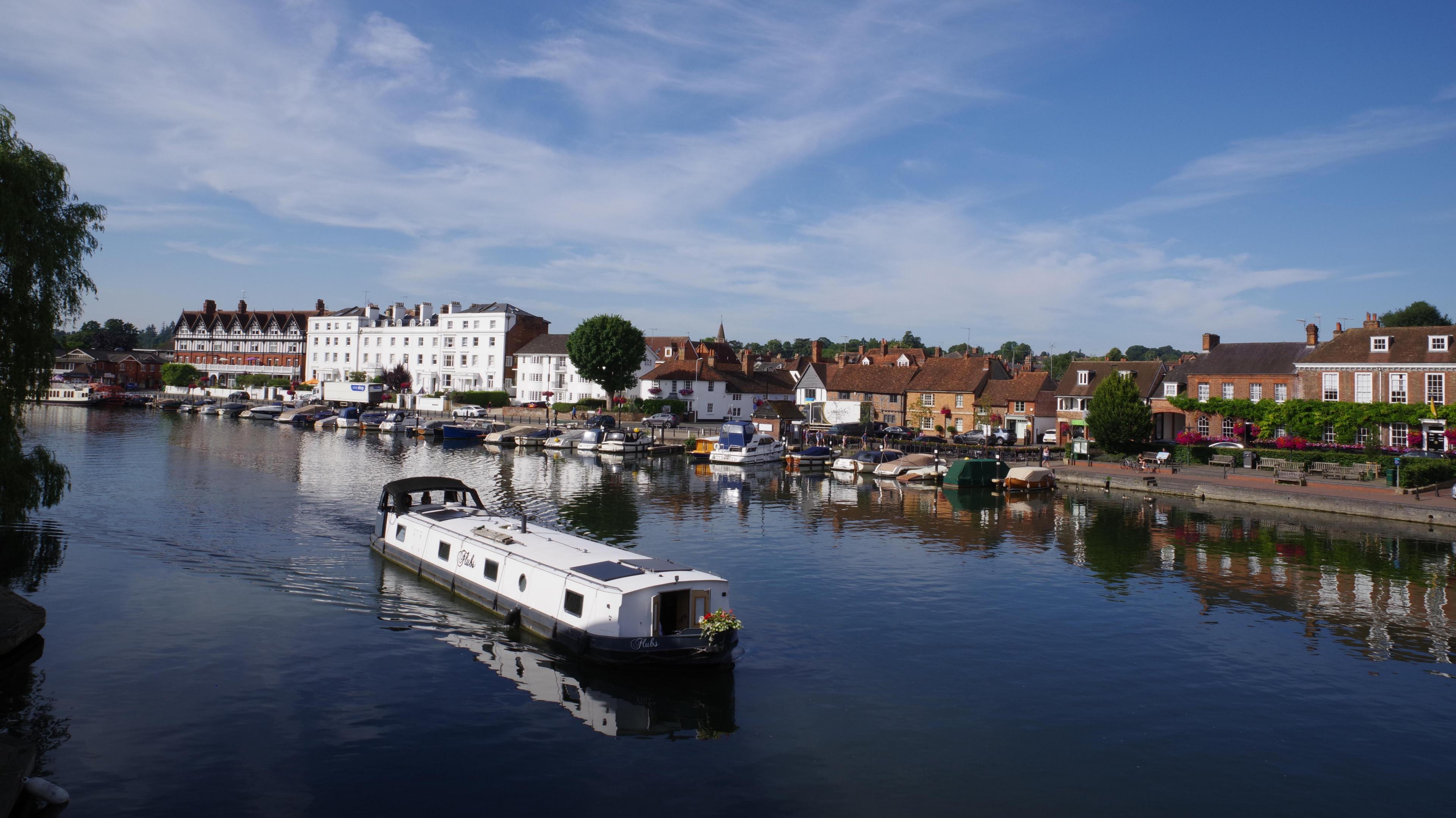 A canal boat sails down a river. On one side of the river are buildings and houses, with smaller boats moored on the bank