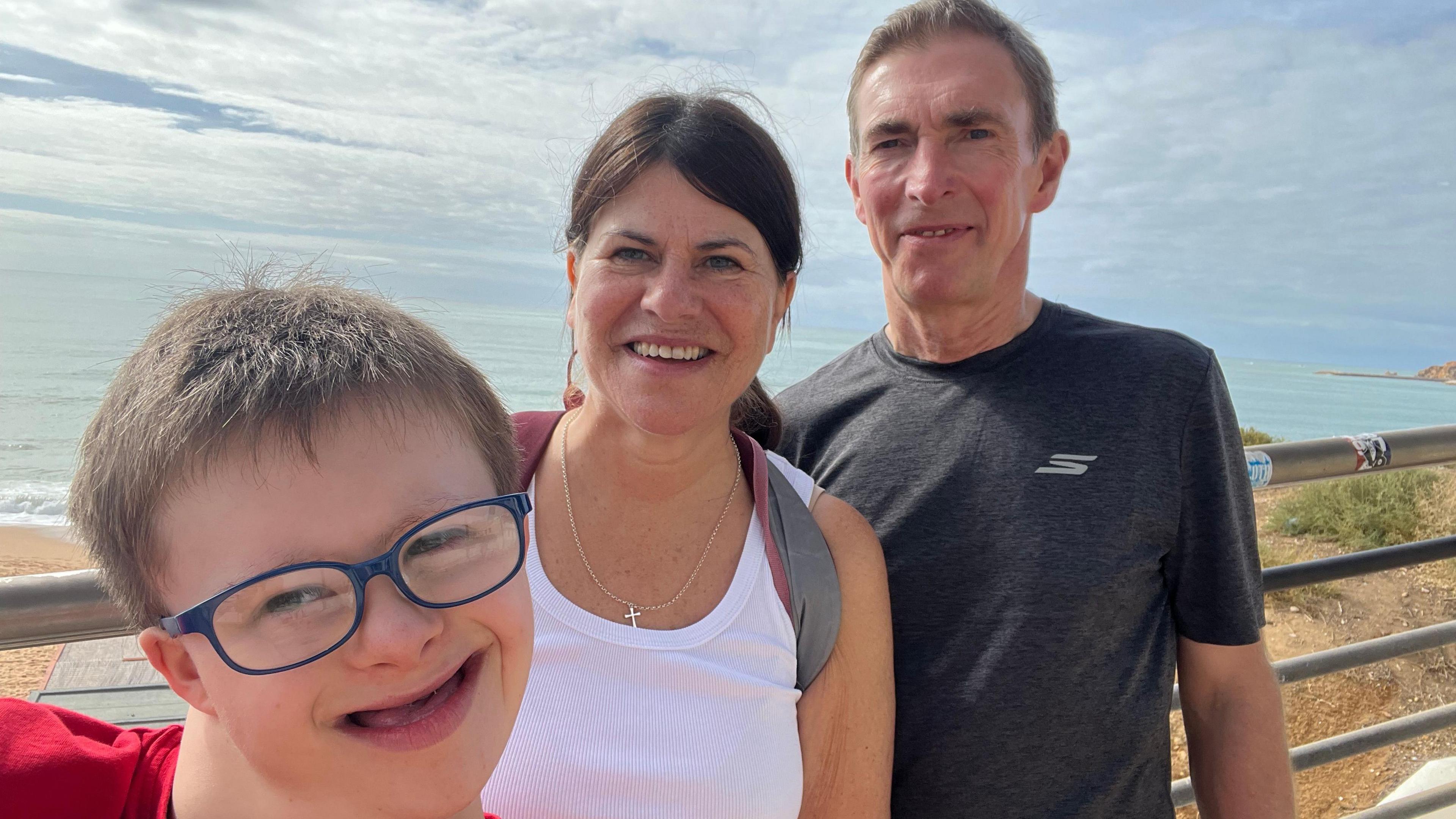 Joe, Anne and Gary, all standing by the sea. Joe is at the front of the picture wearing blue glasses. Anne has on a white T-shirt, with a bag on a her back and Gary has on a black T-shirt. They are by metals railings. 