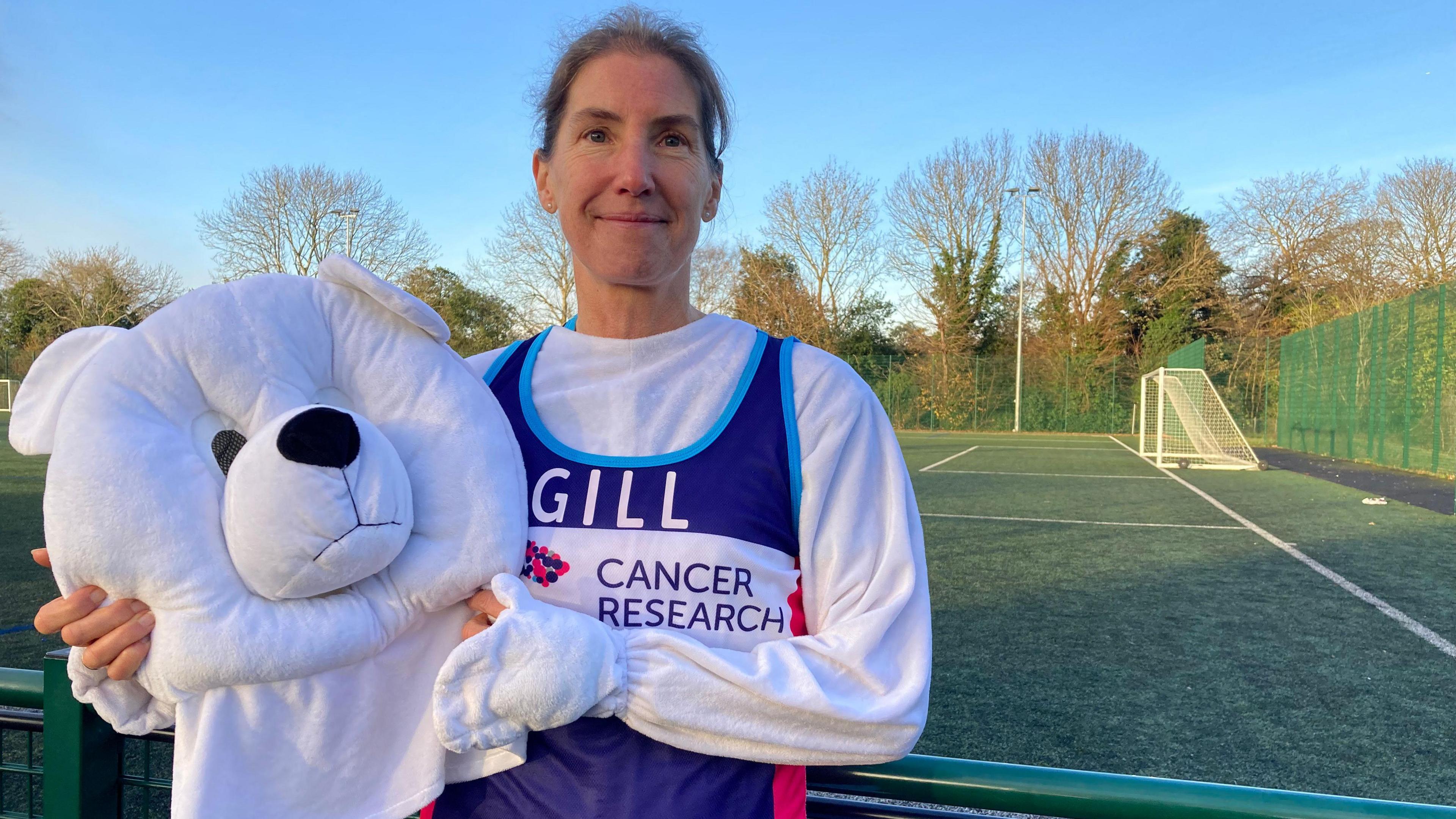 A woman with brown hair in a ponytail, she is smiling while dressed in a white polar bear suit with a purple cancer research bib on that says 'Gill', she is holding the head of the polar bear costume while standing on a football pitch.