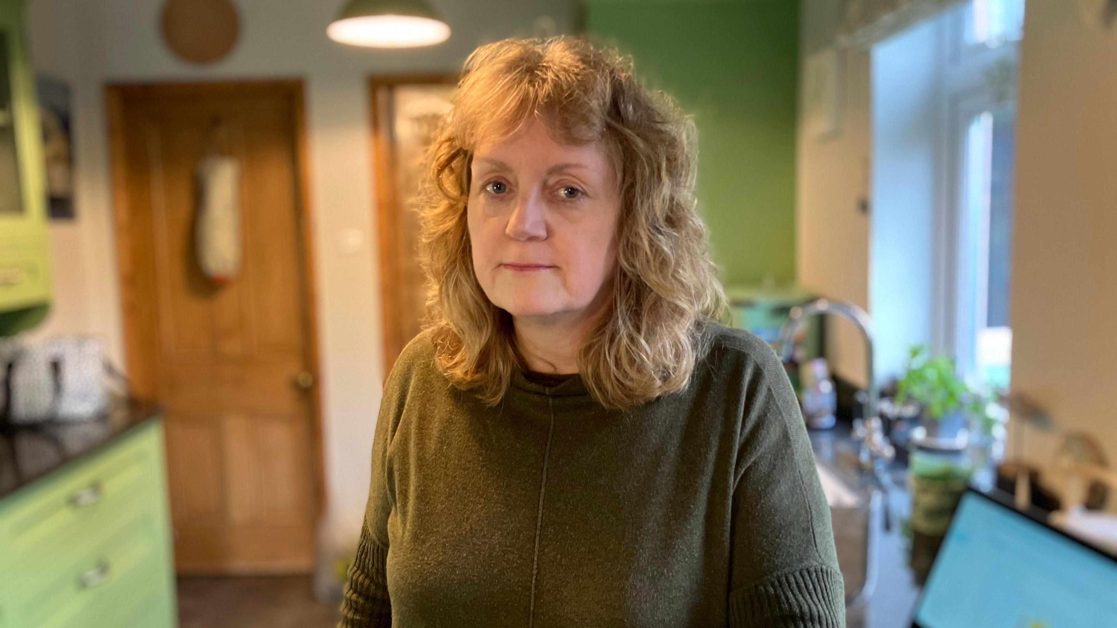 A photograph of the top half of Rachael Clark - she has blonde, curly, shoulder length hair and is wearing a green jumper. She is stood in her kitchen - behind her on the right is a laptop, sink and window. On the back left is a wooden door door and green kitchen cabinets 