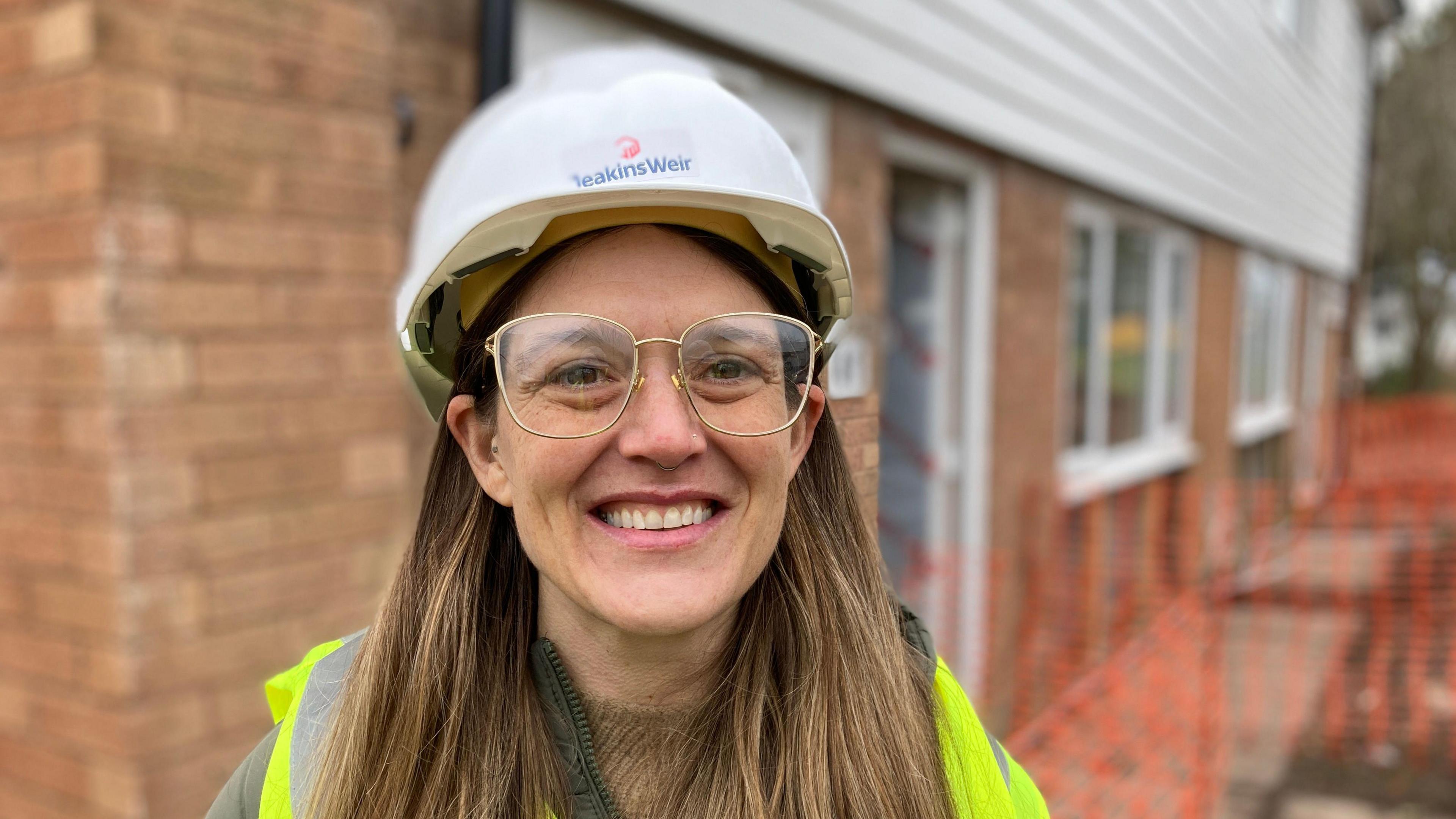 Elly Cutkelvin is a Labour Deputy Mayor at Leicester City Council. She is smiling and wearing glasses and a hard hat, while visiting the construction site on Hospital Close. 