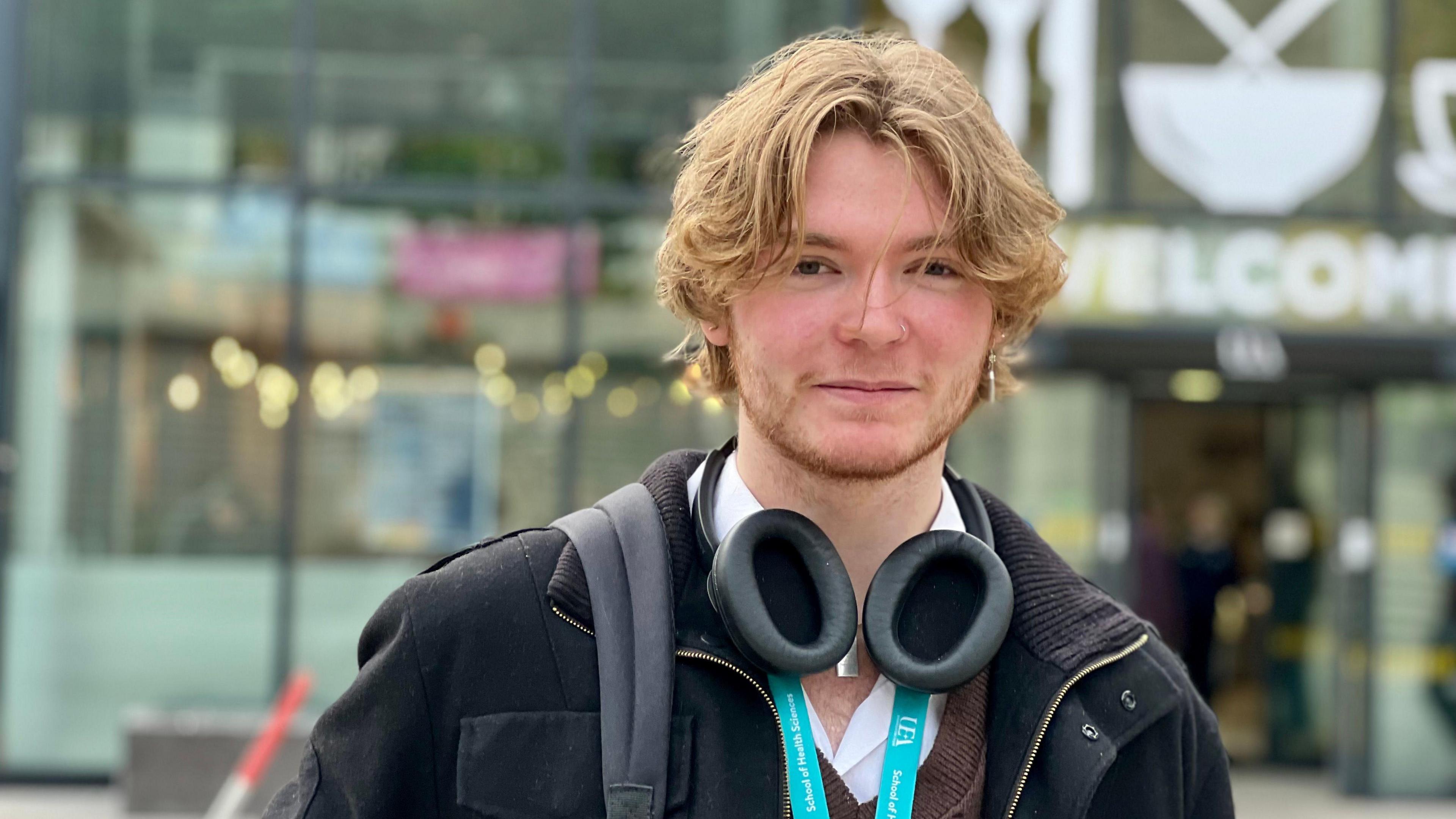 A picture of student, Teddy. He is blond with a beard and is wearing a black coat with his bag slung over his shoulder. 