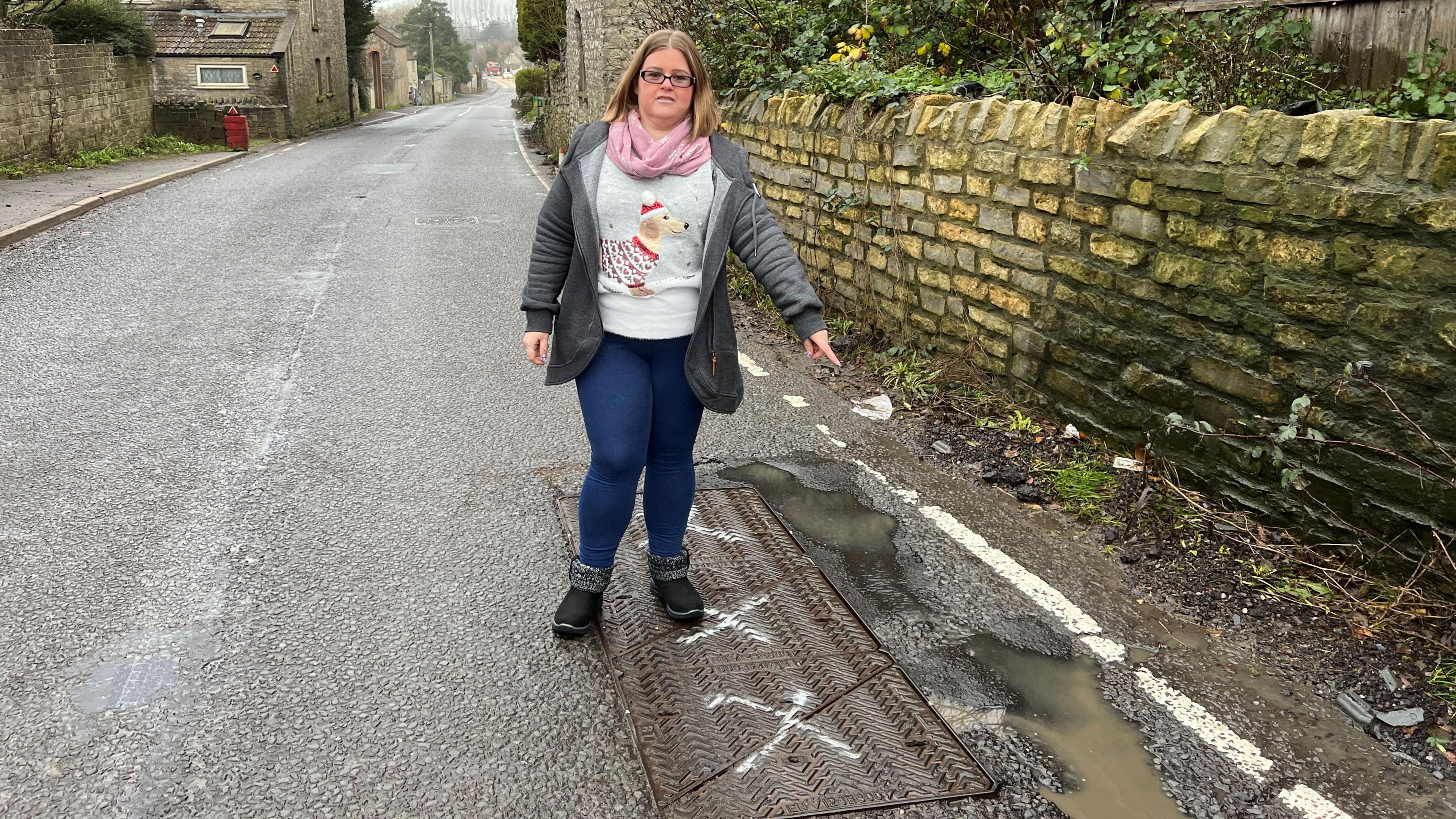 Women stood on a manhole drain cover pointing at a puddle and pothole