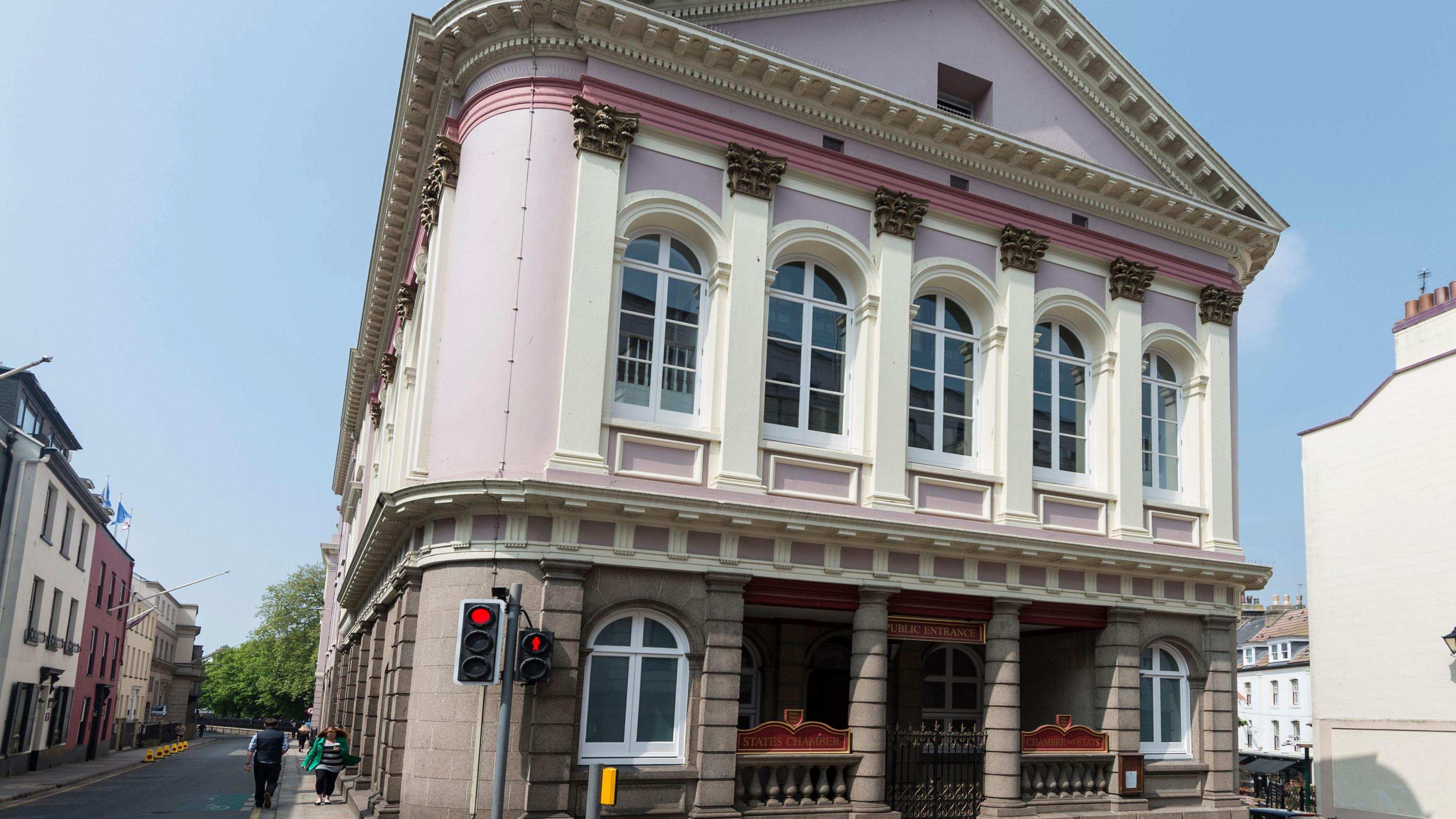 The States Assembly building in Jersey. It has a grey stone lower half and a pink top half with several sash windows. A set of traffic lights is outside the building with its lights on red. People are walking along the pavement next to the building on a sunny day.