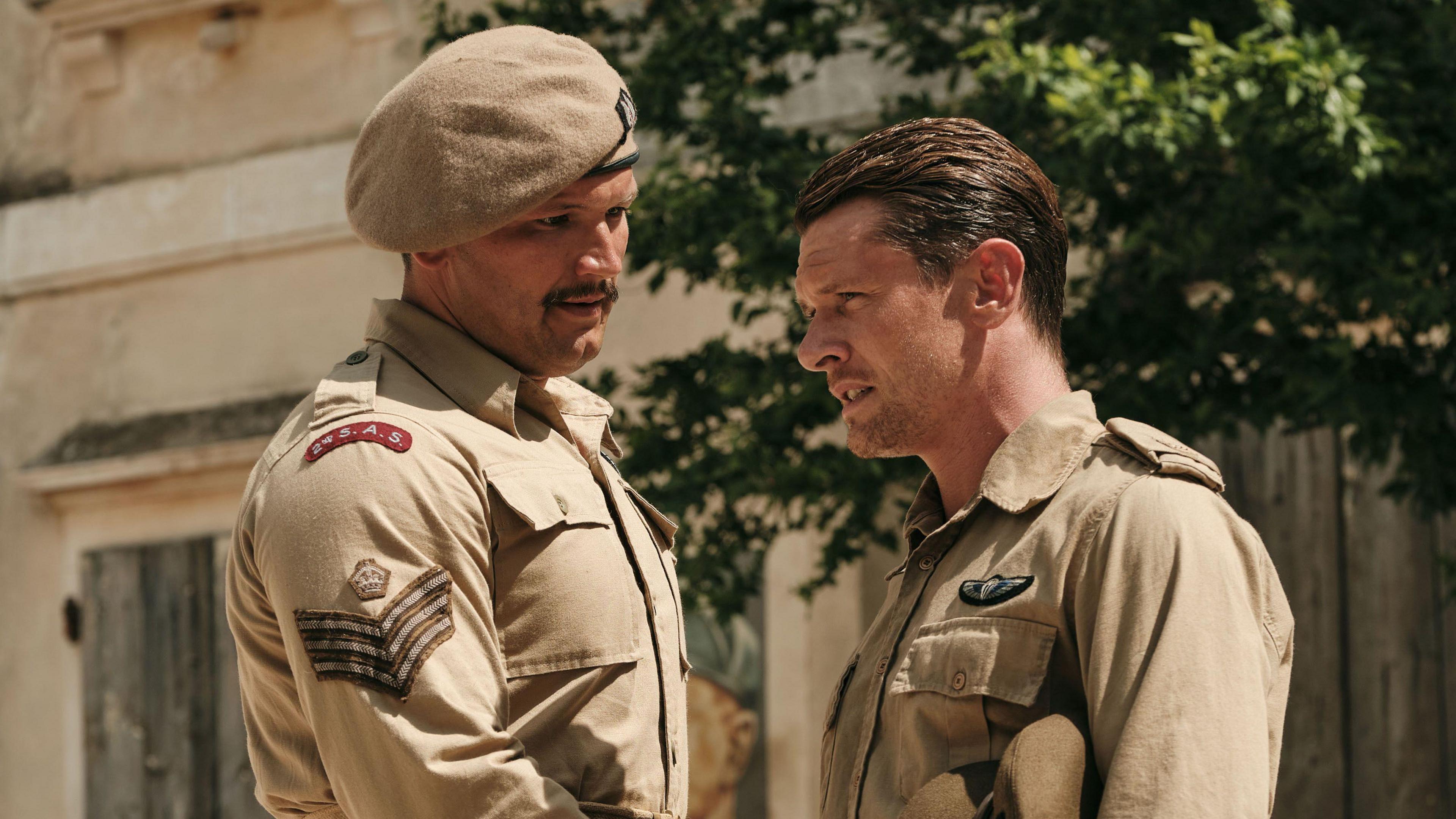 A scene from SAS Rogue Heroes. Corin Silva, who plays Jim Almonds, a large-built man with a moustache, is talking with Jack O'Connell, who plays Paddy Mayne. Both men are wearing khaki uniforms showing SAS insignia.