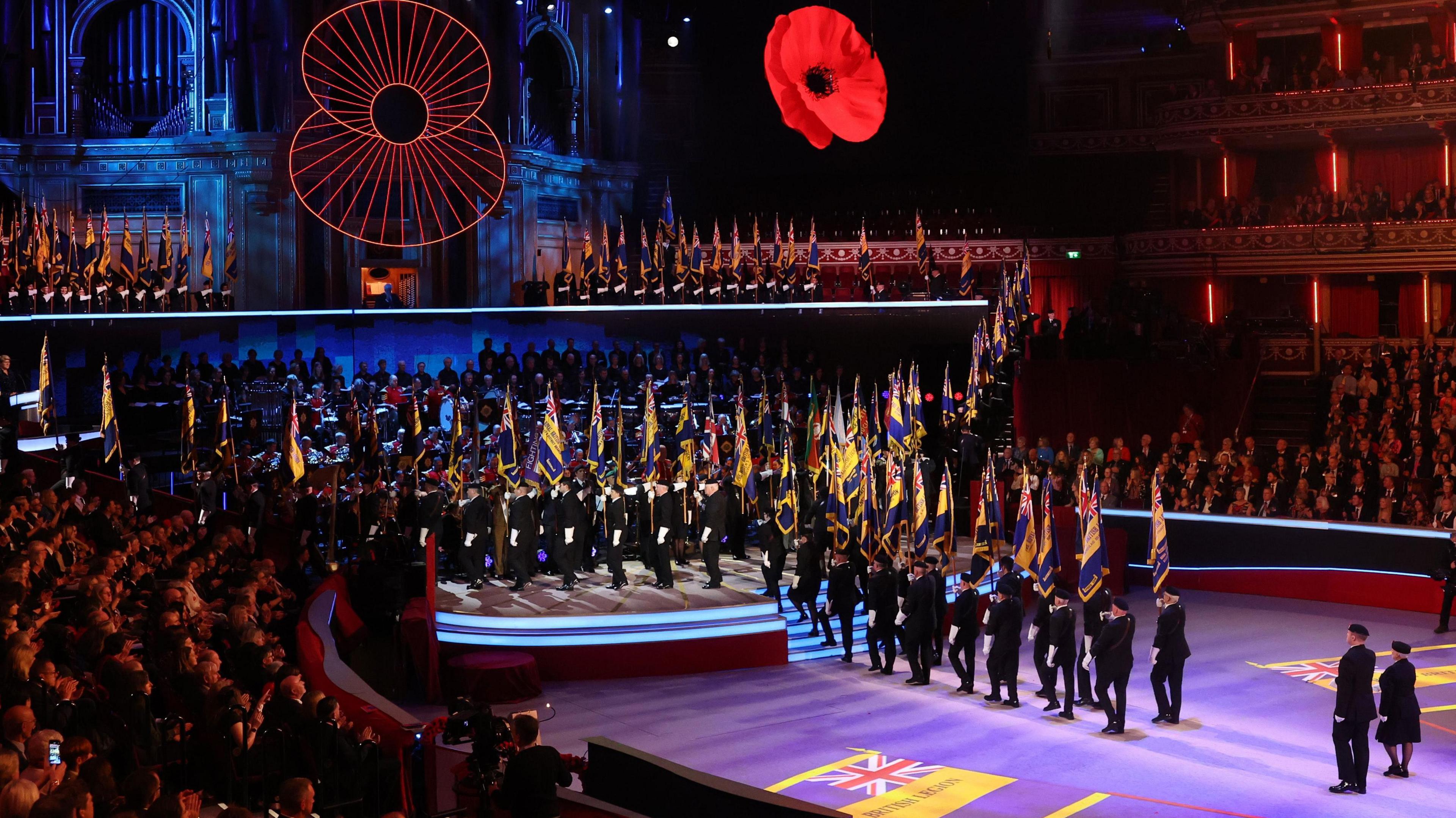 Standard bearers carry flags in the Royal Albert Hall during last year's Festival of Remembrance, with two large poppies above. Hundreds of people watch in the audience.