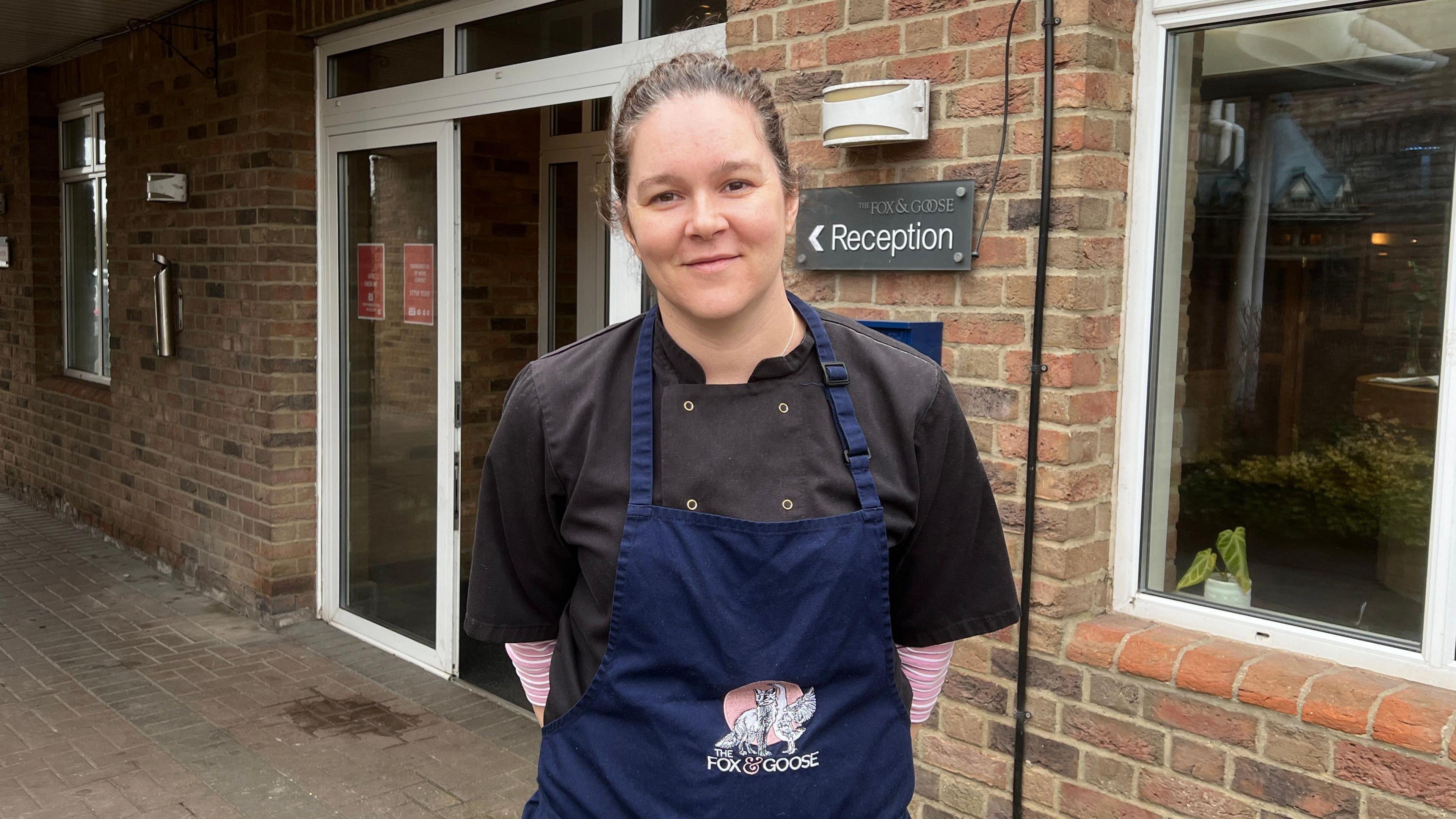 Women stood looking at the camera. She's outside a bricked building. She is wearing an apron saying "the Fox & Goose".