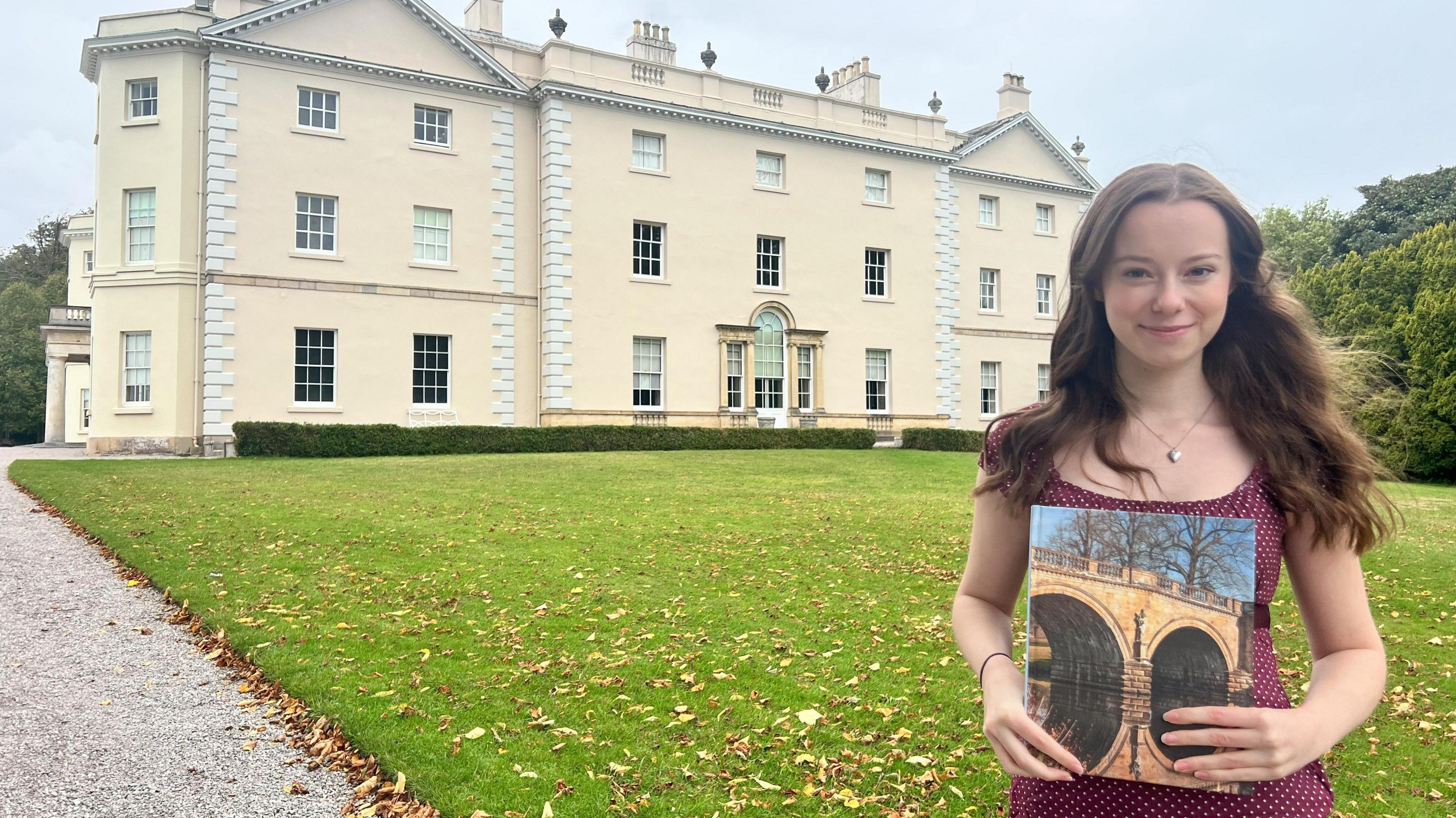A woman looks towards the camera, stood on a patch of grass in front of a large house. She is holding a book with a photograph on the front. The woman has brunette hair and she is wearing a burgundy top.
