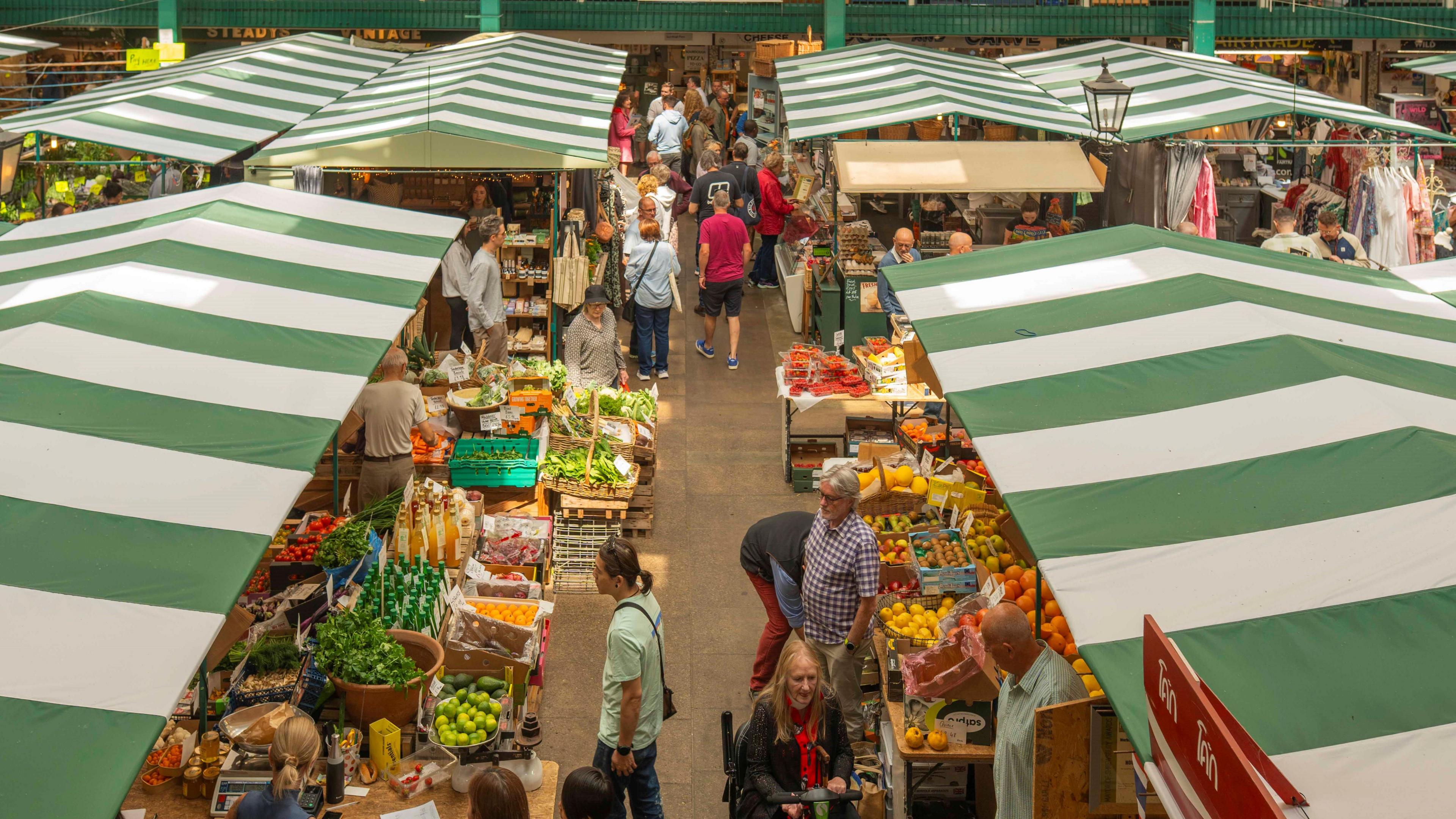 Large, stripey green roof covers stand above the stalls where food, groceries  and clothes can be seen with shoppers walking around them.
