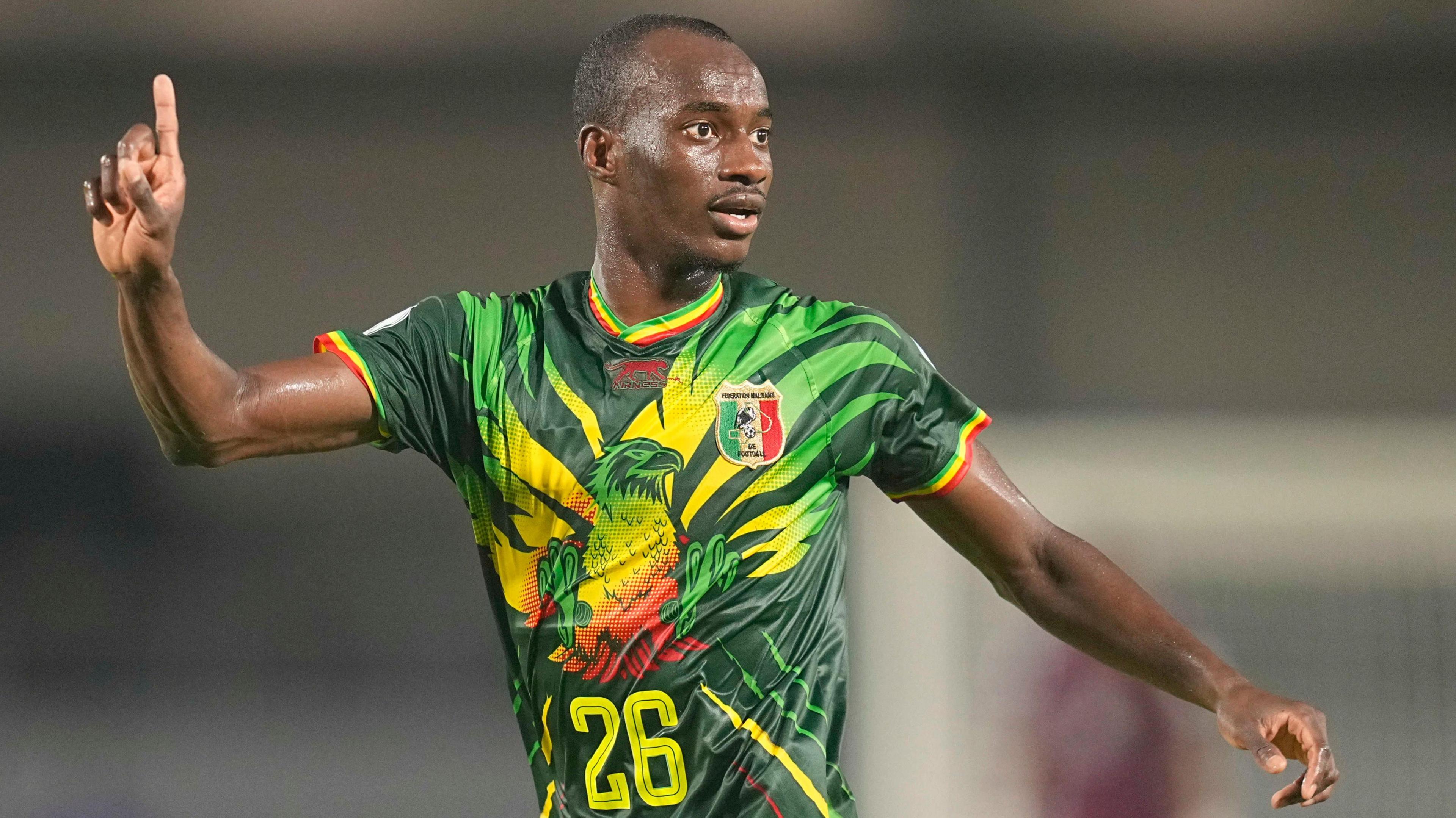 Kamory Doumbia, wearing a green Mali jersey emblazoned with a colourful eagle and the number 26 on the front, points his right index finger in the air as he looks to his left