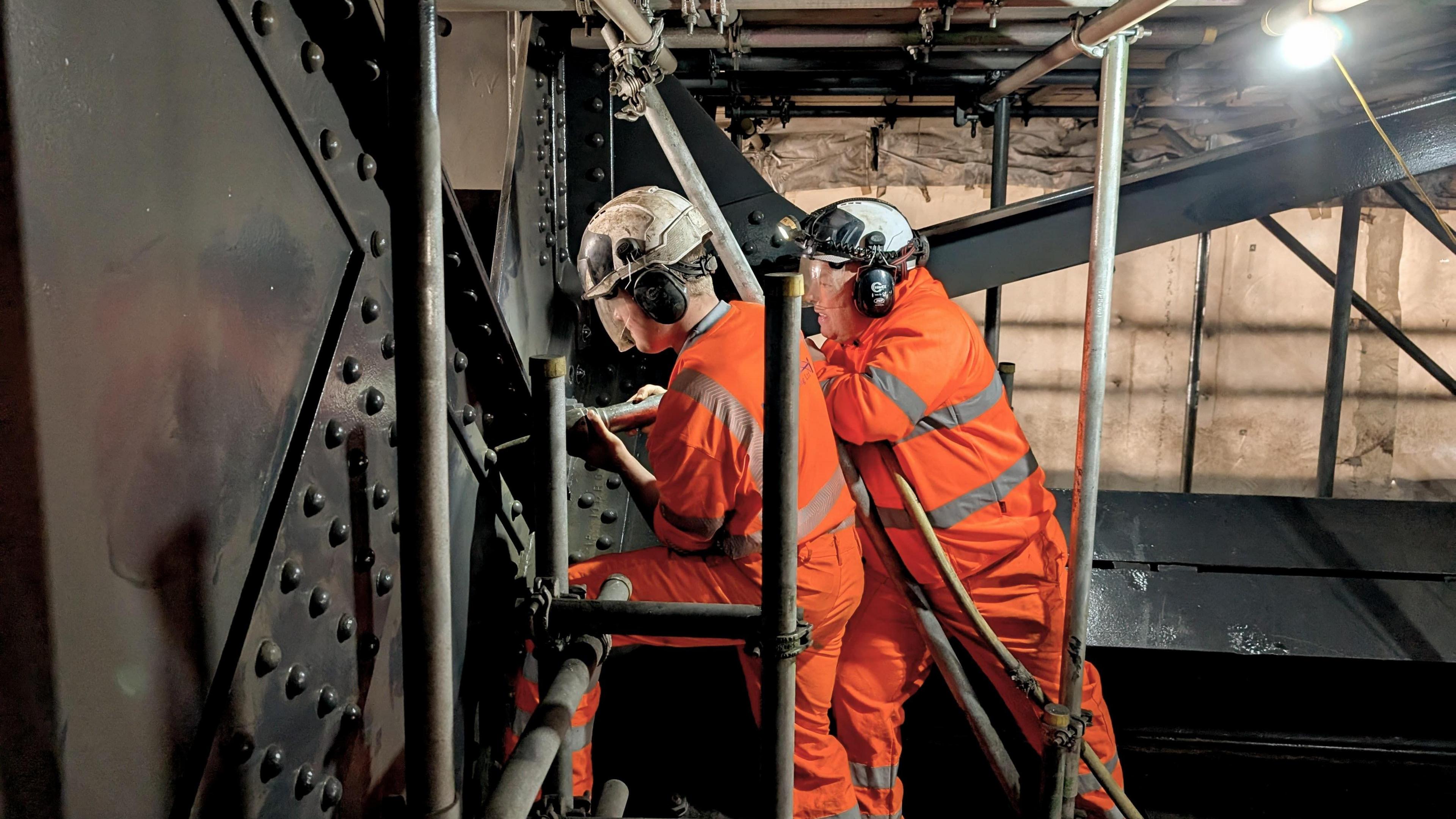 Charlie and Paul hold a pneumatic gun against the rivets on a large wall of steel. They're again surrounded by scaffolding around the other steel beams of the Tyne Bridge. 