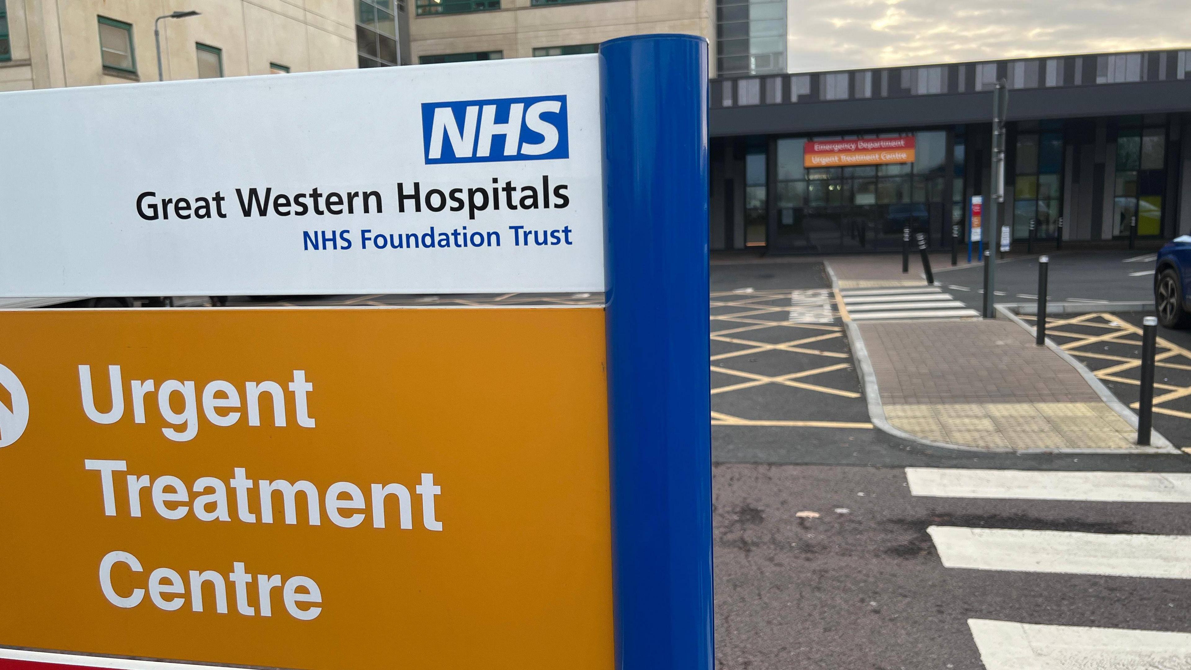 A sign outside a hospital building with the words "NHS Great Western Hospitals NHS Foundation Trust" and a second sign on an orange background below with the words "Urgent treatment centre". Hospital buildings and zebra crossings on the right.