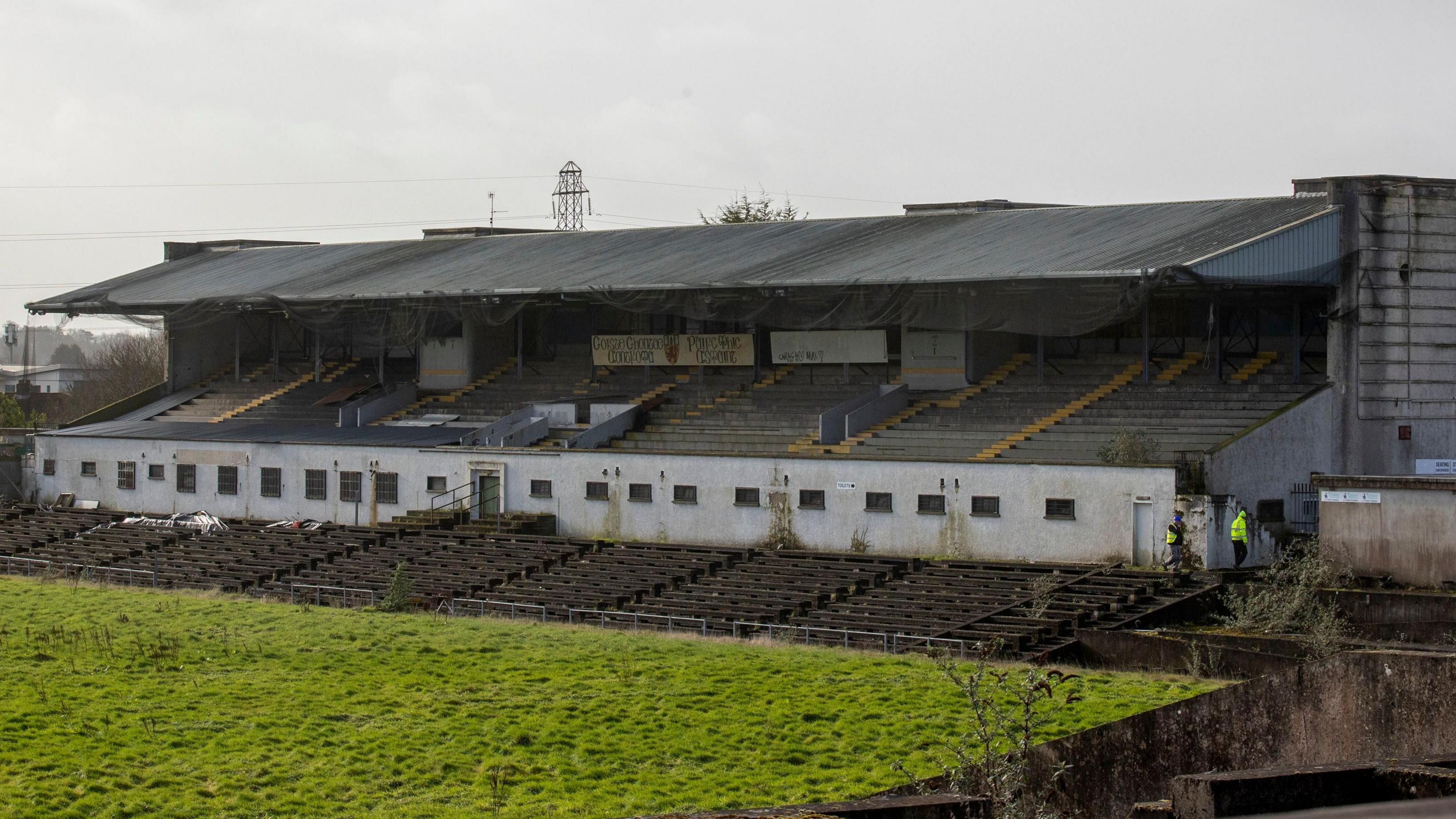 A wide shot of the disused Casement Park stadium in west Belfast. The grass is overgrown and the seats are empty. In the distance, two workmen in hi-vis jackets can be seen walking around the complex.