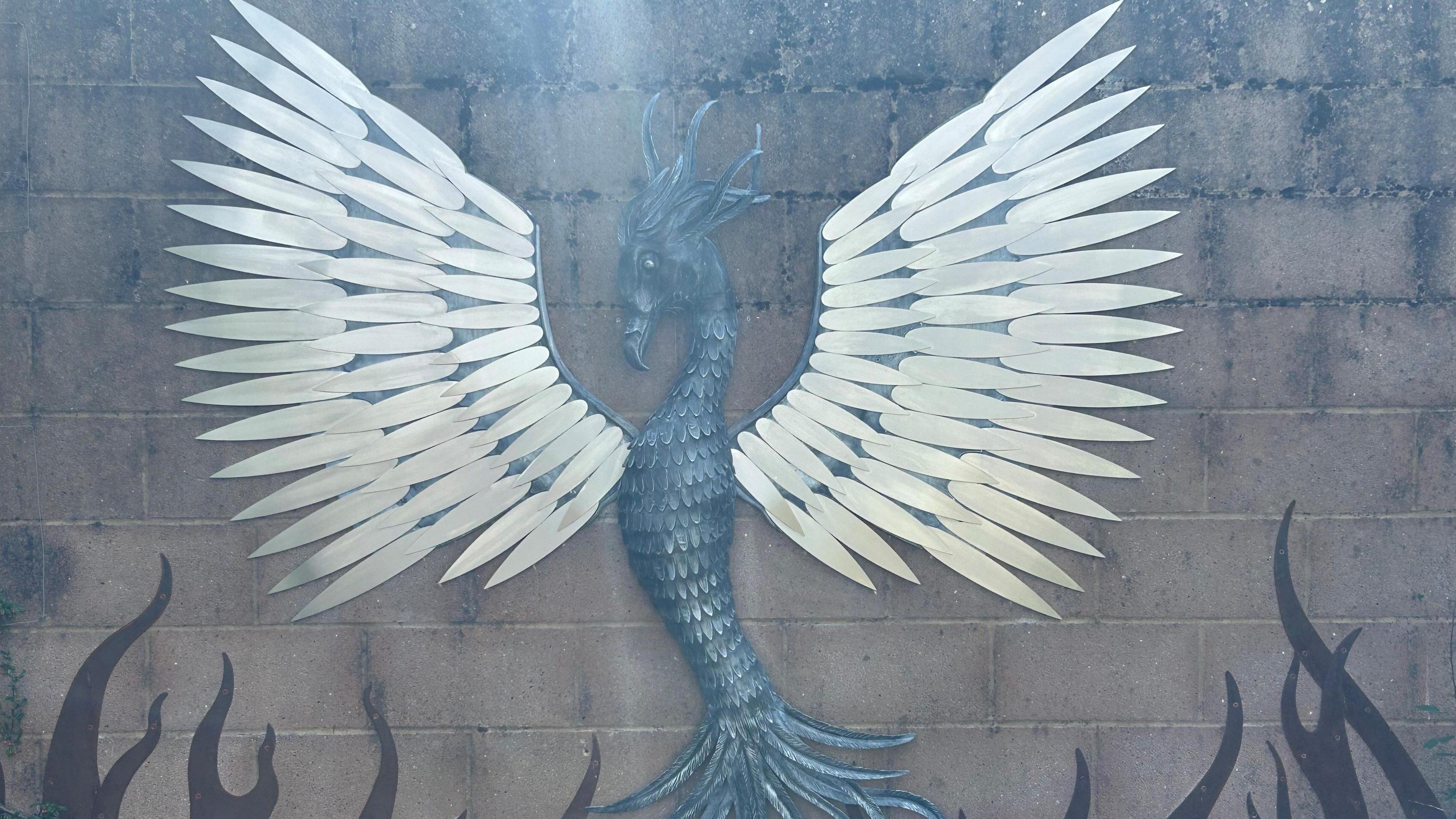 A wider view of the full steel phoenix sculpture. It has outstretched wings above a bed of flames. The feathers that make up its wings are made of three sizes, growing larger the further out on the wing they are.