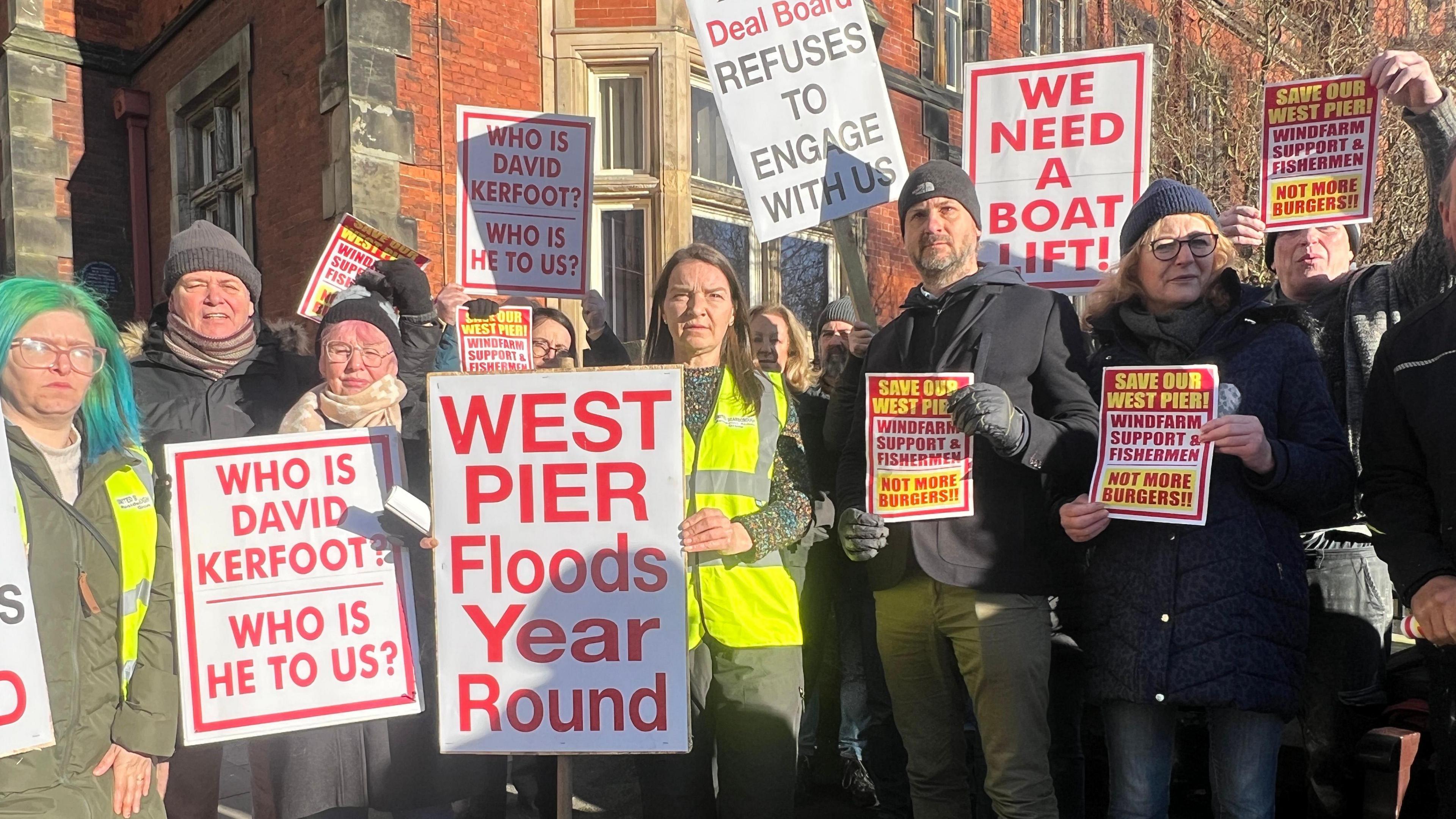 Men and women stand with placards that read "Who is David Kerfoot?", "we need a boat lift" and "West Pier floods year round".