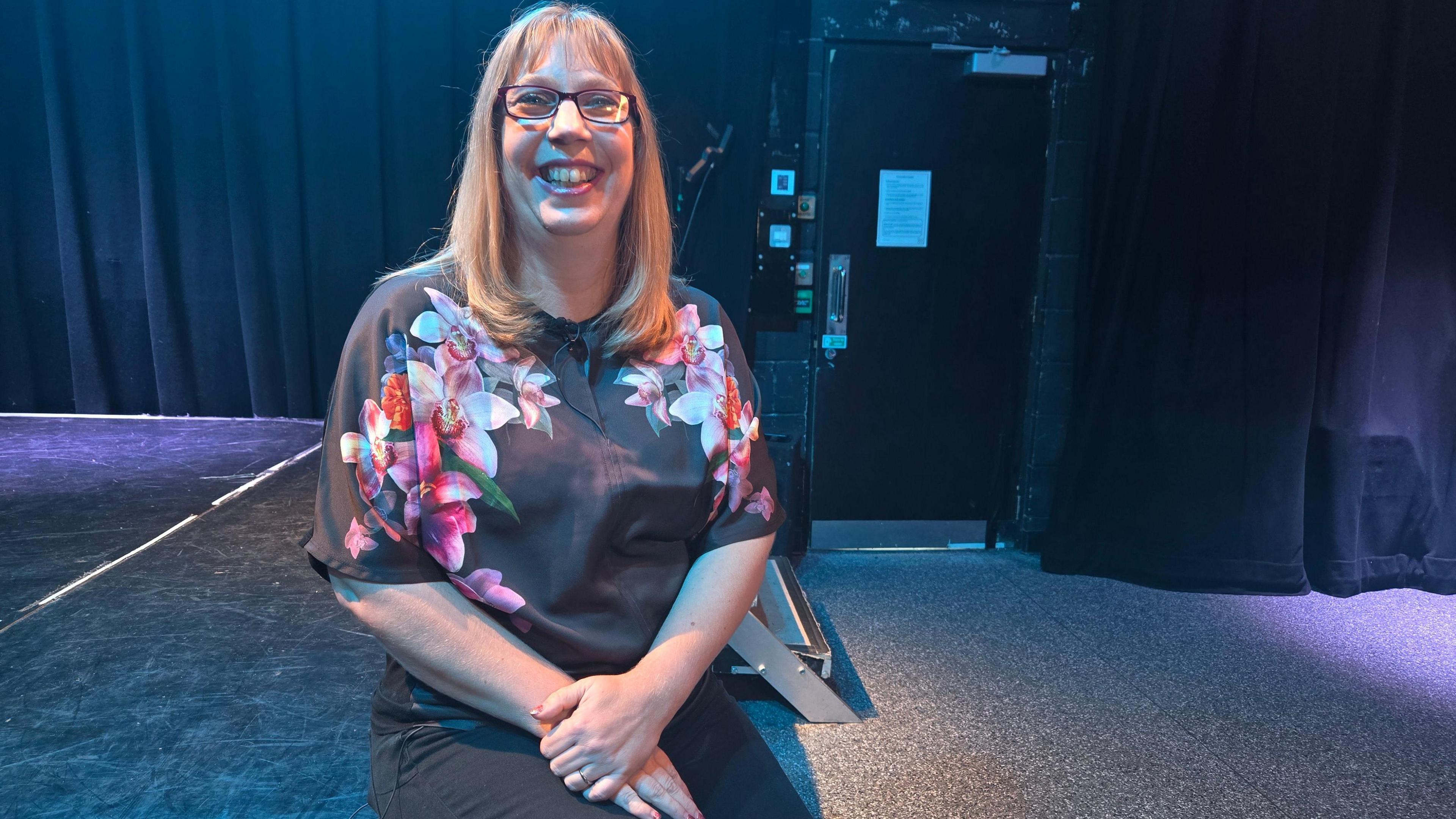 Zoe Wickham sitting on the edge of a stage in a performance hall or theatre. She has her hand on one knee and is smiling at the camera. She has long blonde hair.