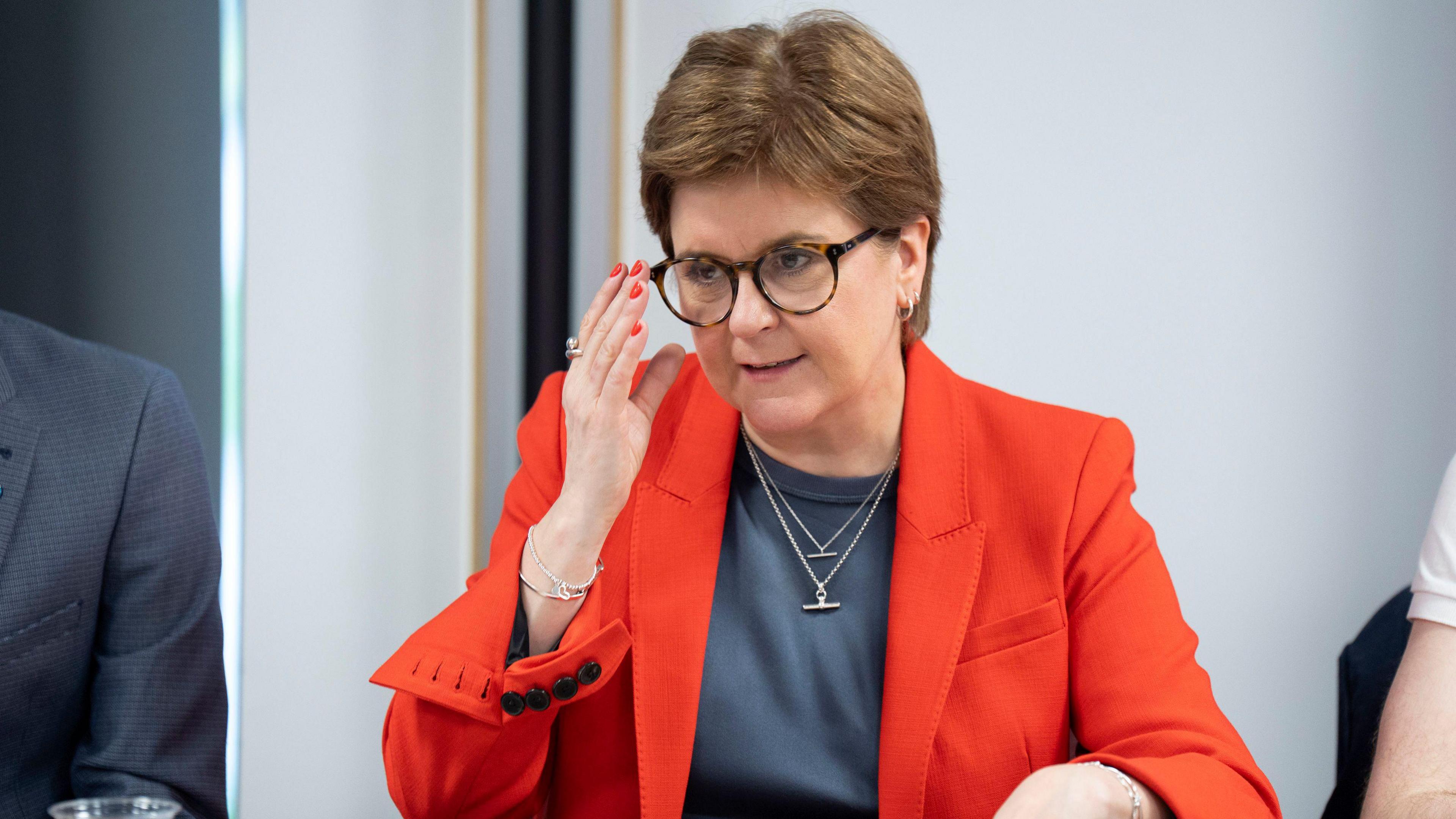 Nicola Sturgeon wears glasses and a red blazer and looks focused