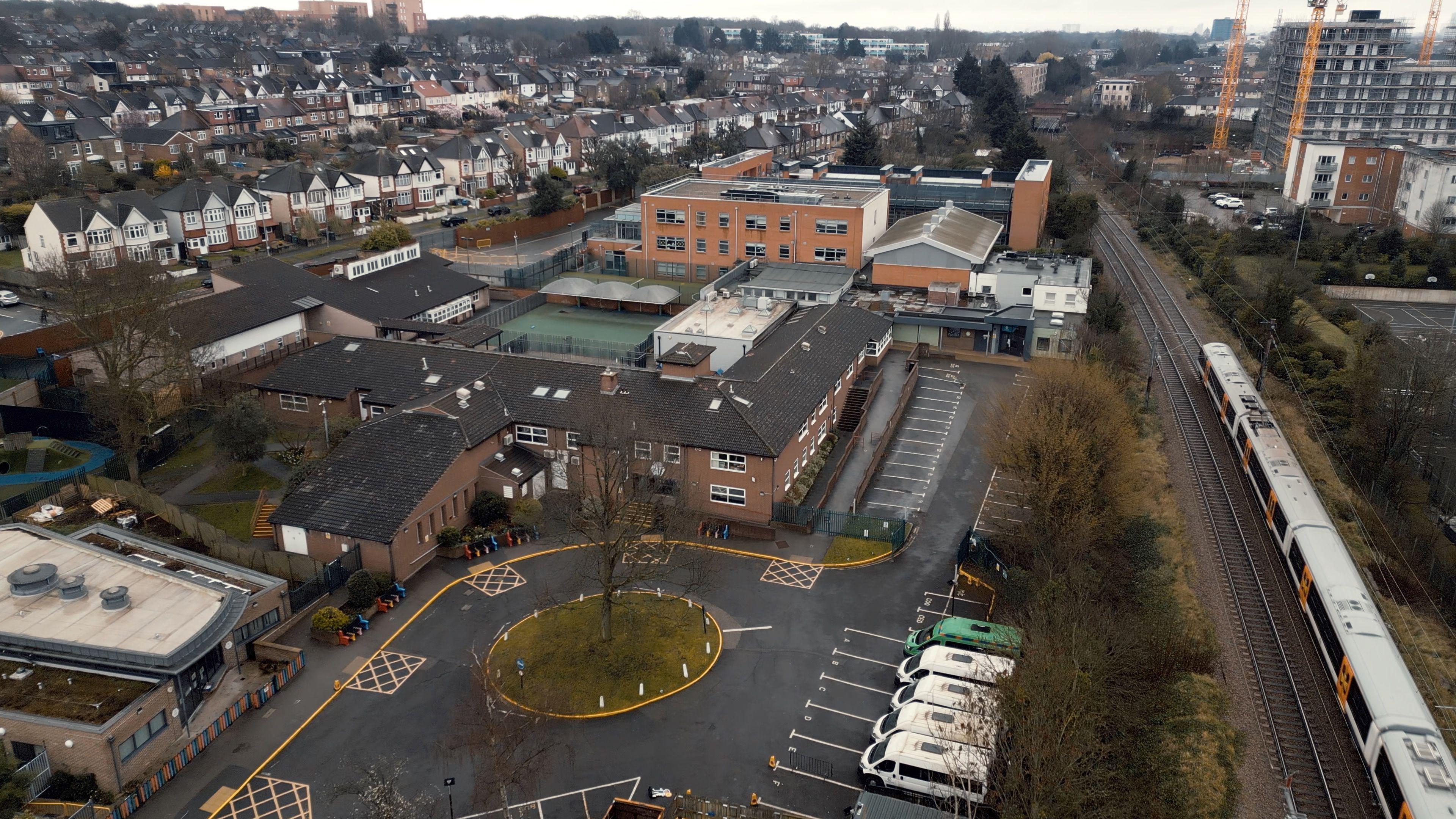 An aerial view of Whitefield School in north-east London, showing a two-storey red-brick building with a more modern extension at the rear. A London Overground line runs next to the site, with new tower blocks beyond it, while on the other side are streets of semi-detached homes.