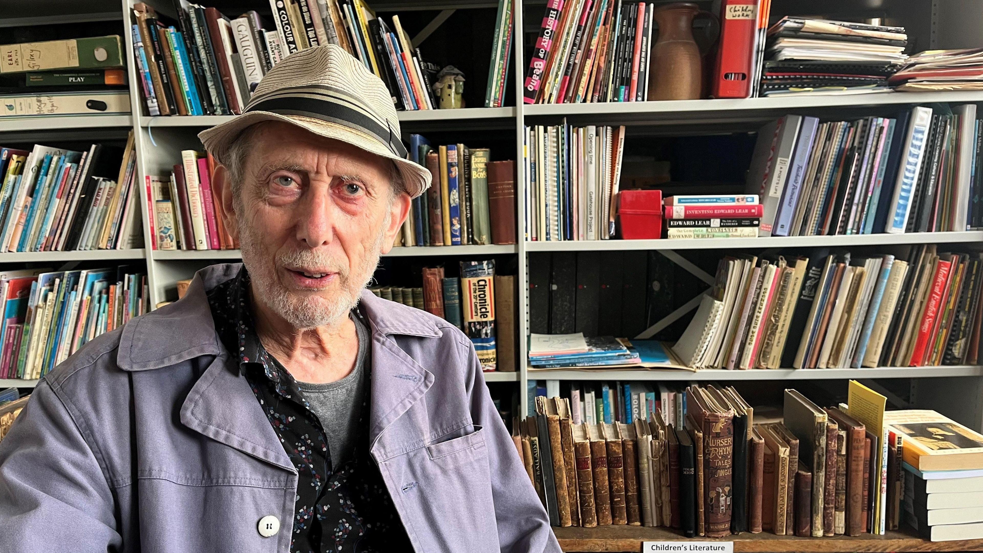 Michael Rosen is pictured in his office left of frame, with shelves of books behind him. He wears a white hat with a dark band around it and a lilac jacket.