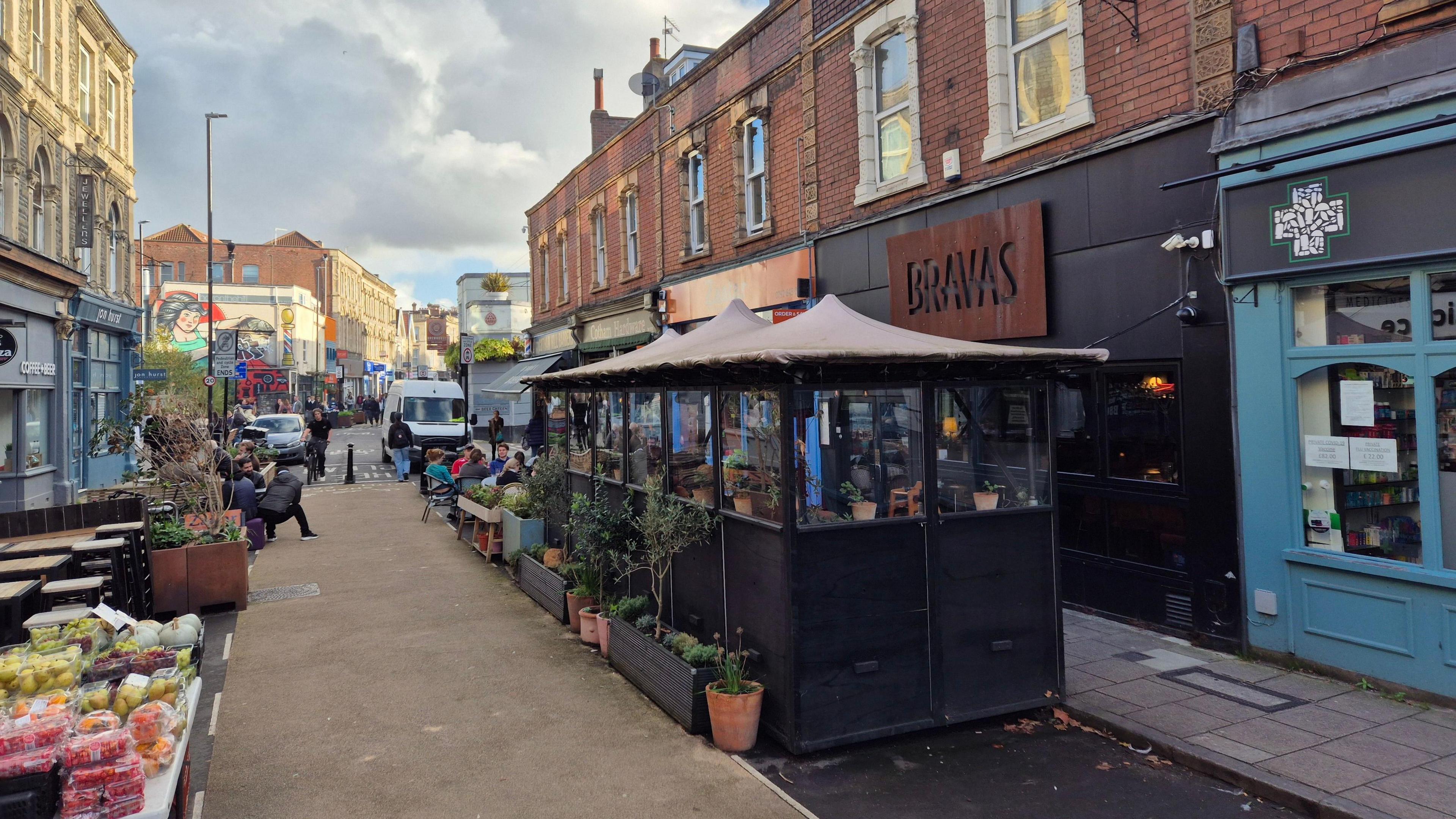 An image of Bravas with the black outdoor seating with brown roof covering. To the left of the structure, you can see the pedestrianised street with restaurants on the other side. There is a table of fruit and veg in the bottom left of the photo. The rust-coloured Bravas sign is above the restaurant.