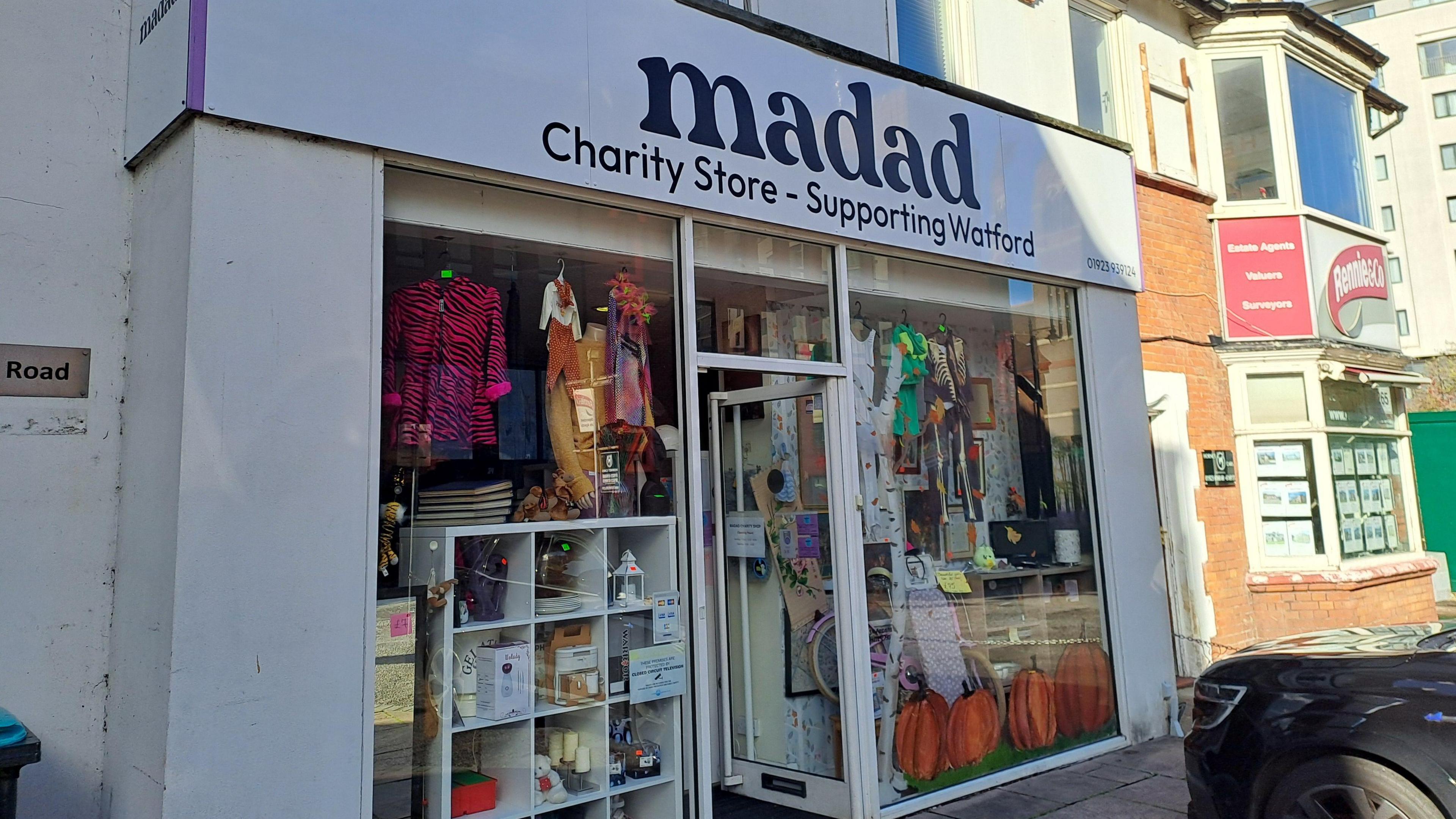 A charity shop named “madad” is shown, with a sign reading “Charity Store – Supporting Watford”. The storefront has large glass windows displaying various items, including colourful clothing, accessories, and household goods. 