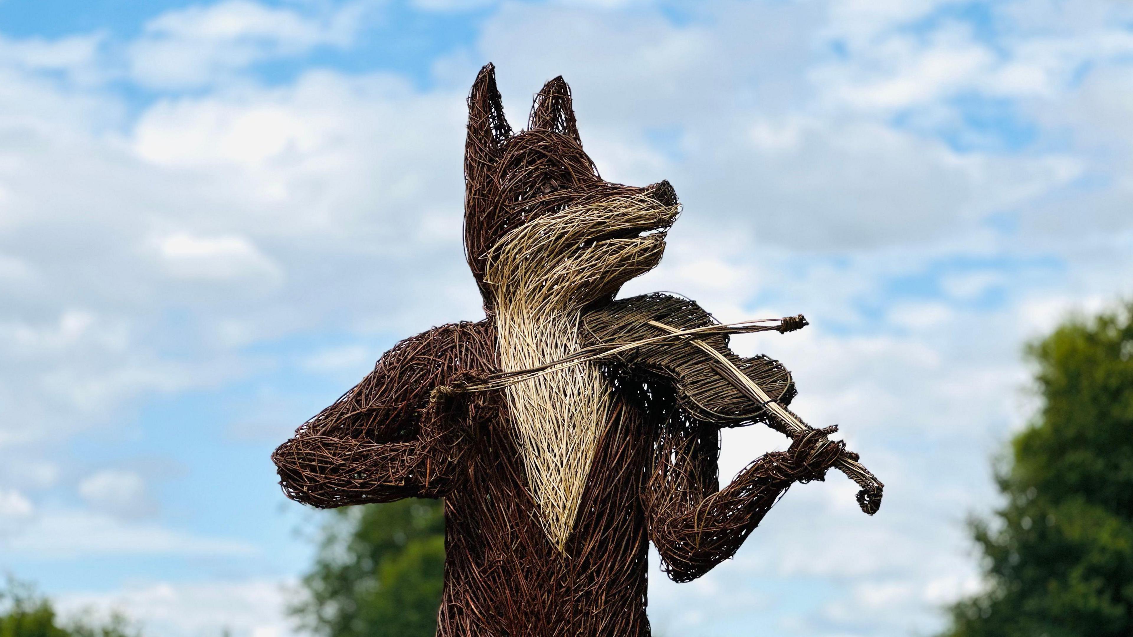 A large sculpture made out of twigs resembles a fox playing a fiddle. It stands in the field at the 2024 folk festival in Cambridge. It is a bright and dry sunny day with blue sky and a few white clouds. 