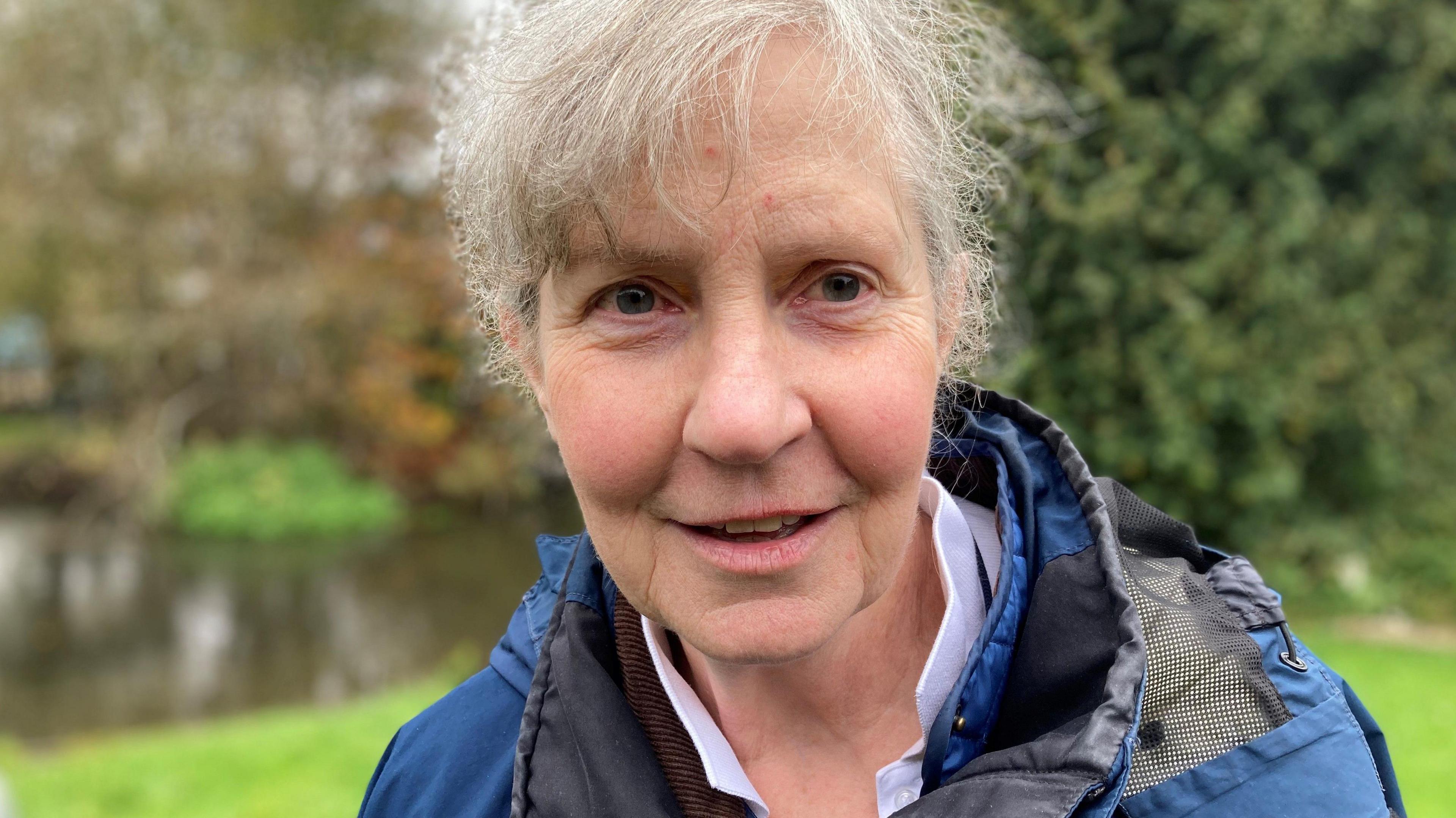 Angela Jordan smiling at the camera, has grey hair and is wearing a blue waterproof jacket, with greenery and the River Lambourn in the background.