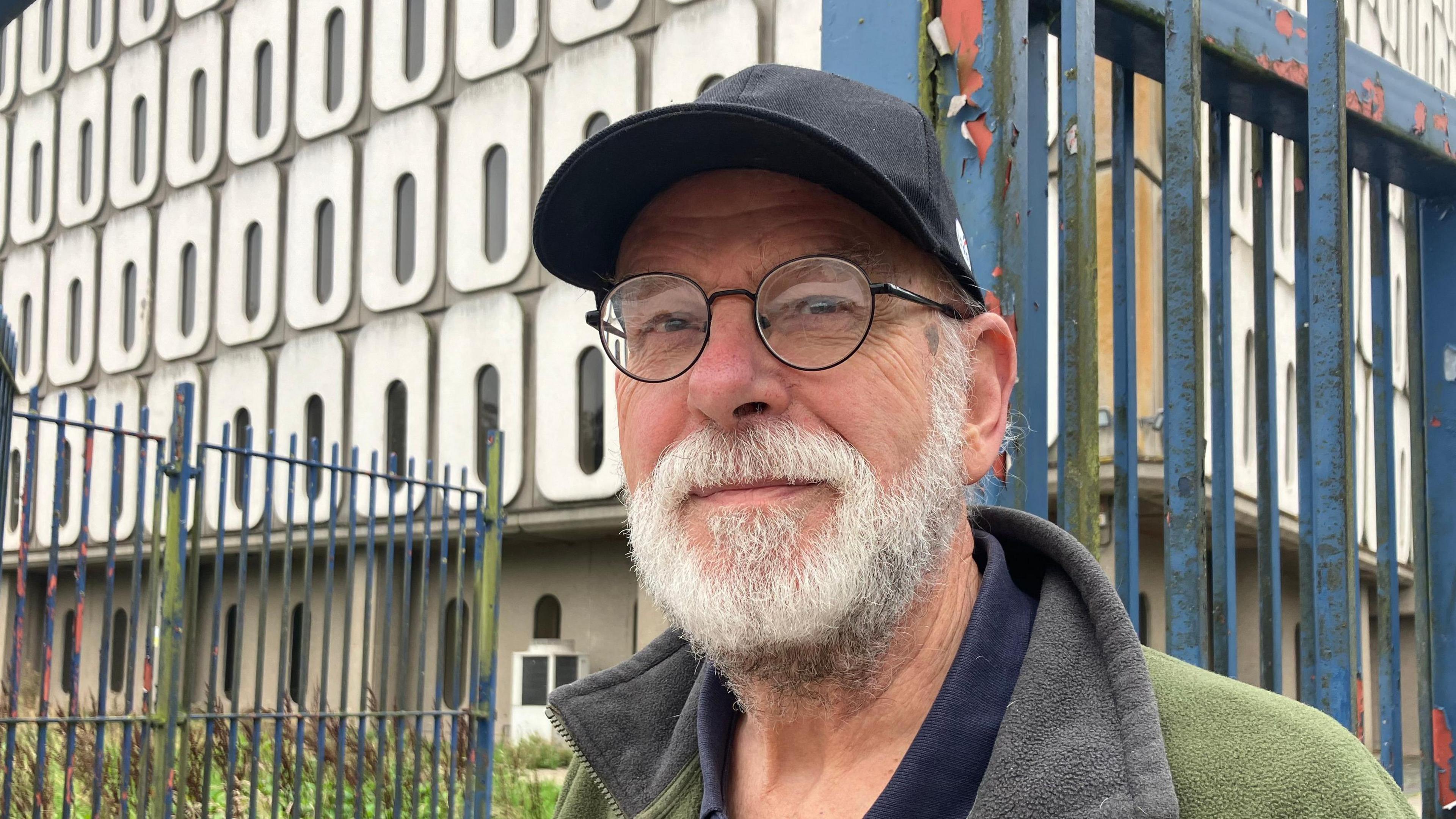 John stands smiling outside Marlborough House, he is wearing a black cap and has a white beard. He is also wearing black framed glasses and a green fleece coat.