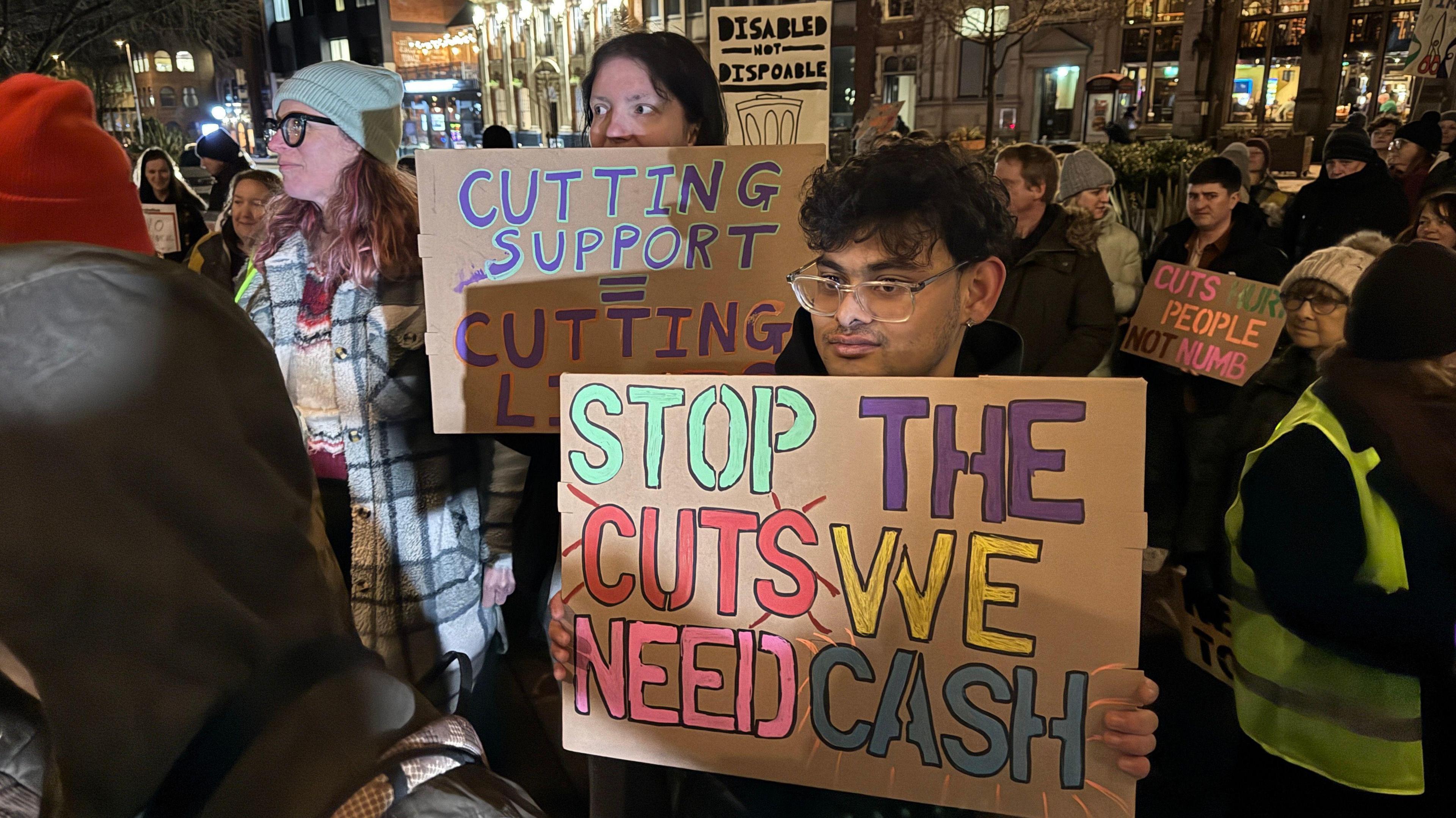 Protesters hold colourful placards outside. The one in the foreground is held by a person in glasses and reads 'stop the cuts we need cash'.