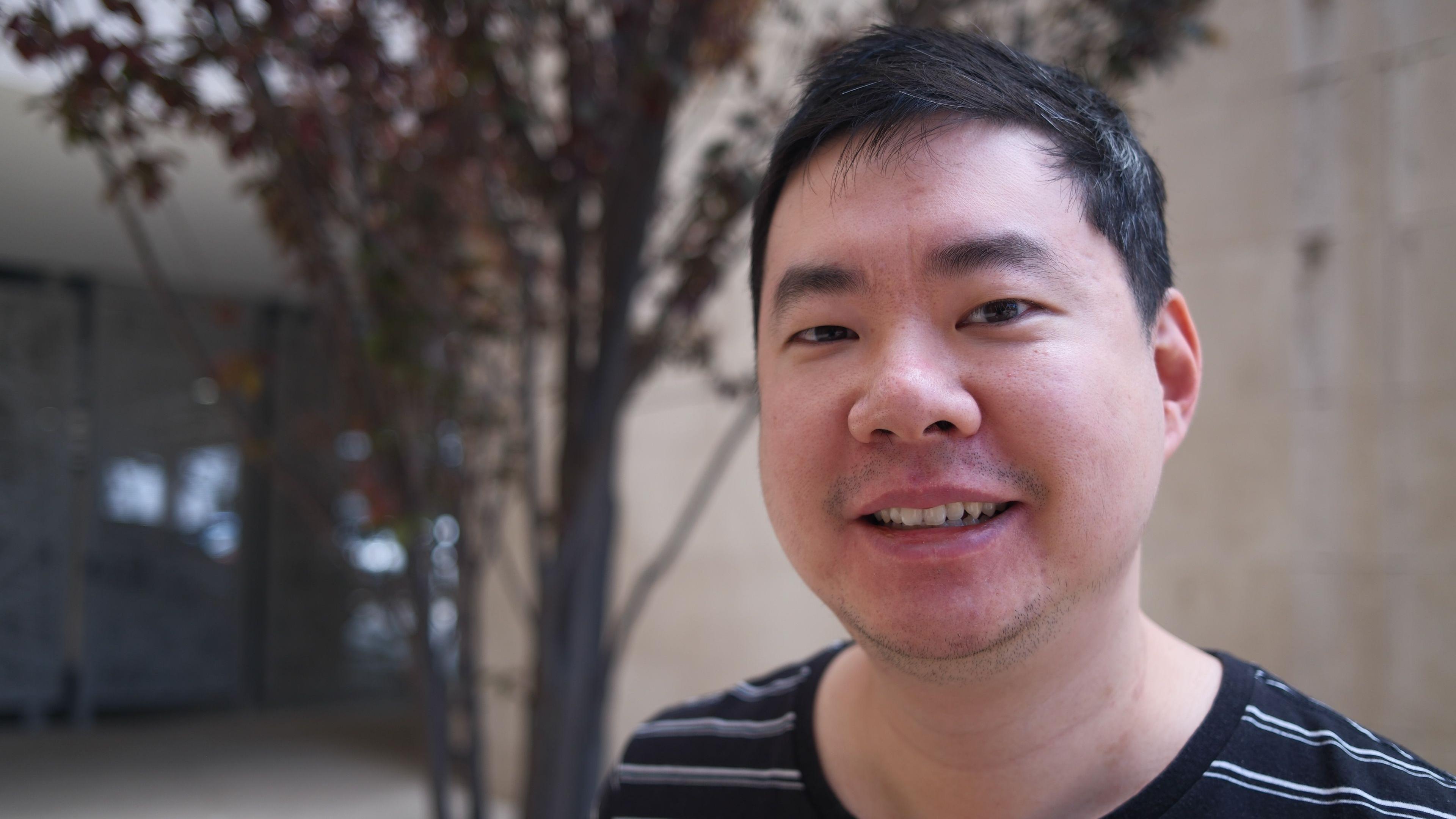 A man with short dark hair standing in front of a building and a tree smiles at the camera.