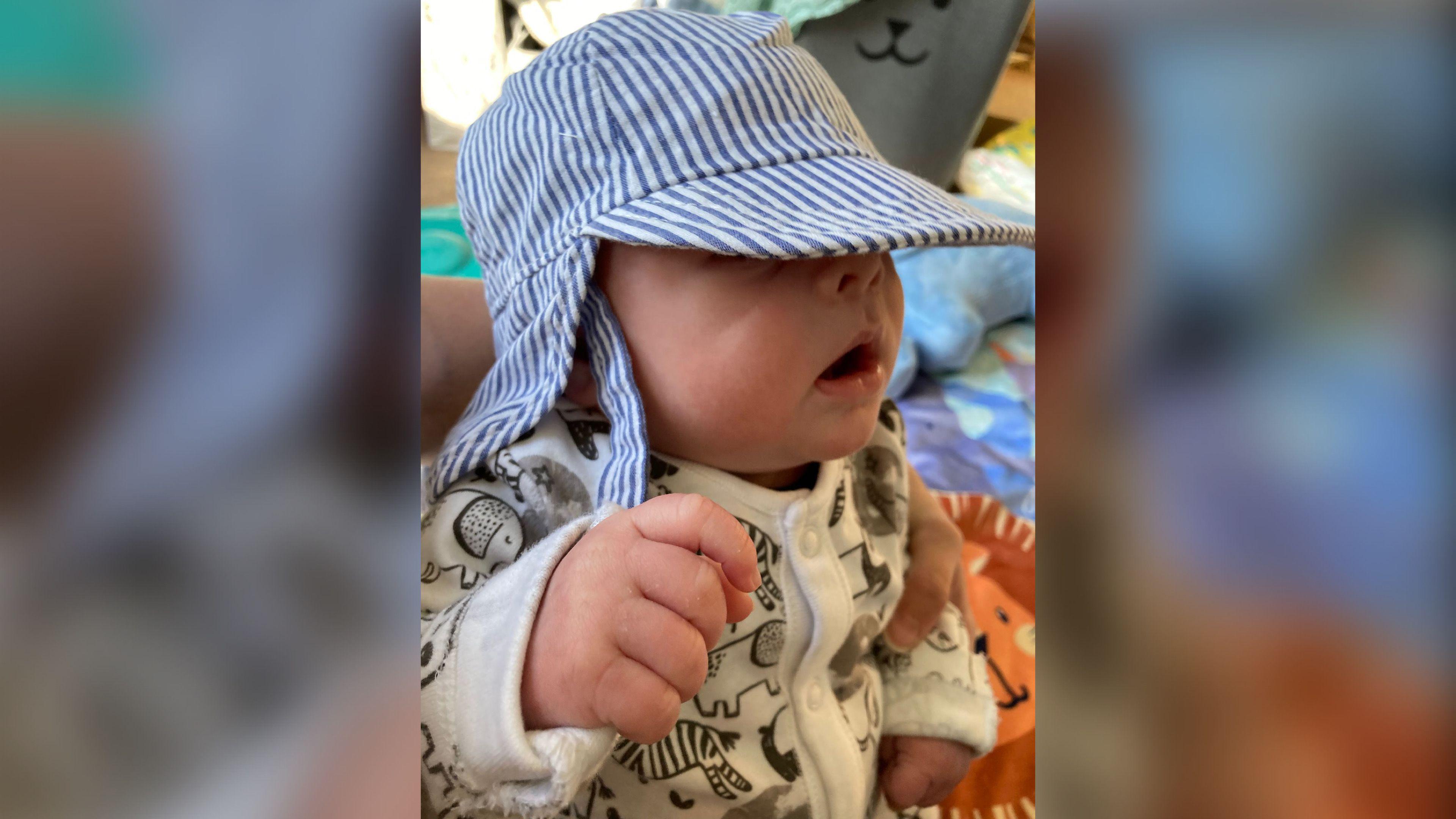 Gina's newborn son wearing a striped blue and grey son hat and a white baby gro which has black cartoon pictures of animals on